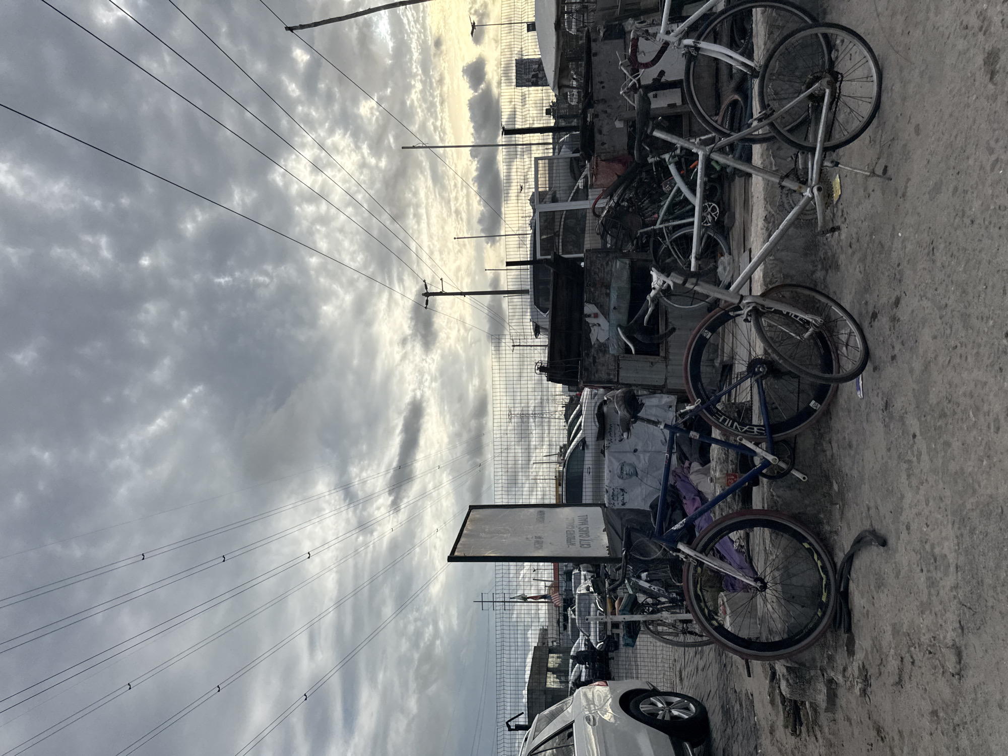 The image shows a street scene in what appears to be an urban or suburban area, possibly Igboefon, Nigeria. The focus is on a collection of bicycles leaning against a wall or structure on the right side of the frame. These bikes appear to be a mix of styles and conditions, some seemingly abandoned or stored.
Behind the bicycles is a fence, and beyond that, there are parked cars and possibly a building or structure. A sign is visible near the bicycles, but the text is illegible. Overhead, there are power lines stretching across the sky, which is overcast with cloudy skies. The pavement is visible in the foreground. No people are distinctly visible in the image.