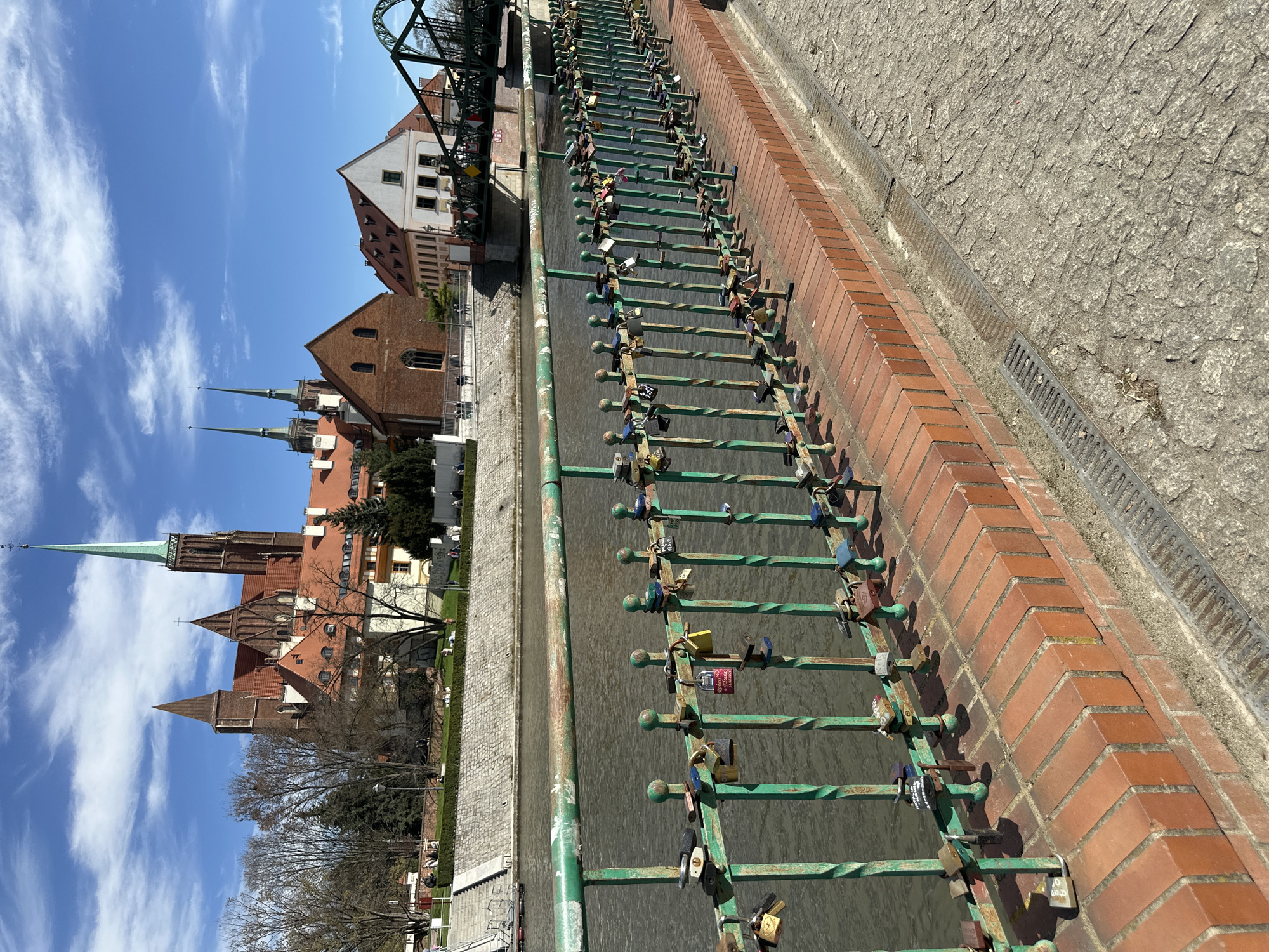 The image shows a canal in Wrocław, Poland. A green metal fence runs along a brick wall on one side of the canal, and many padlocks, some old and rusted, are attached to the fence. The canal water is murky. Beyond the canal, there are buildings with red roofs, including the spires of a church or cathedral. A green metal bridge is visible in the upper part of the image. The sky is blue with wispy clouds. On the upper right side of the frame a gray cobblestone pavement is seen.