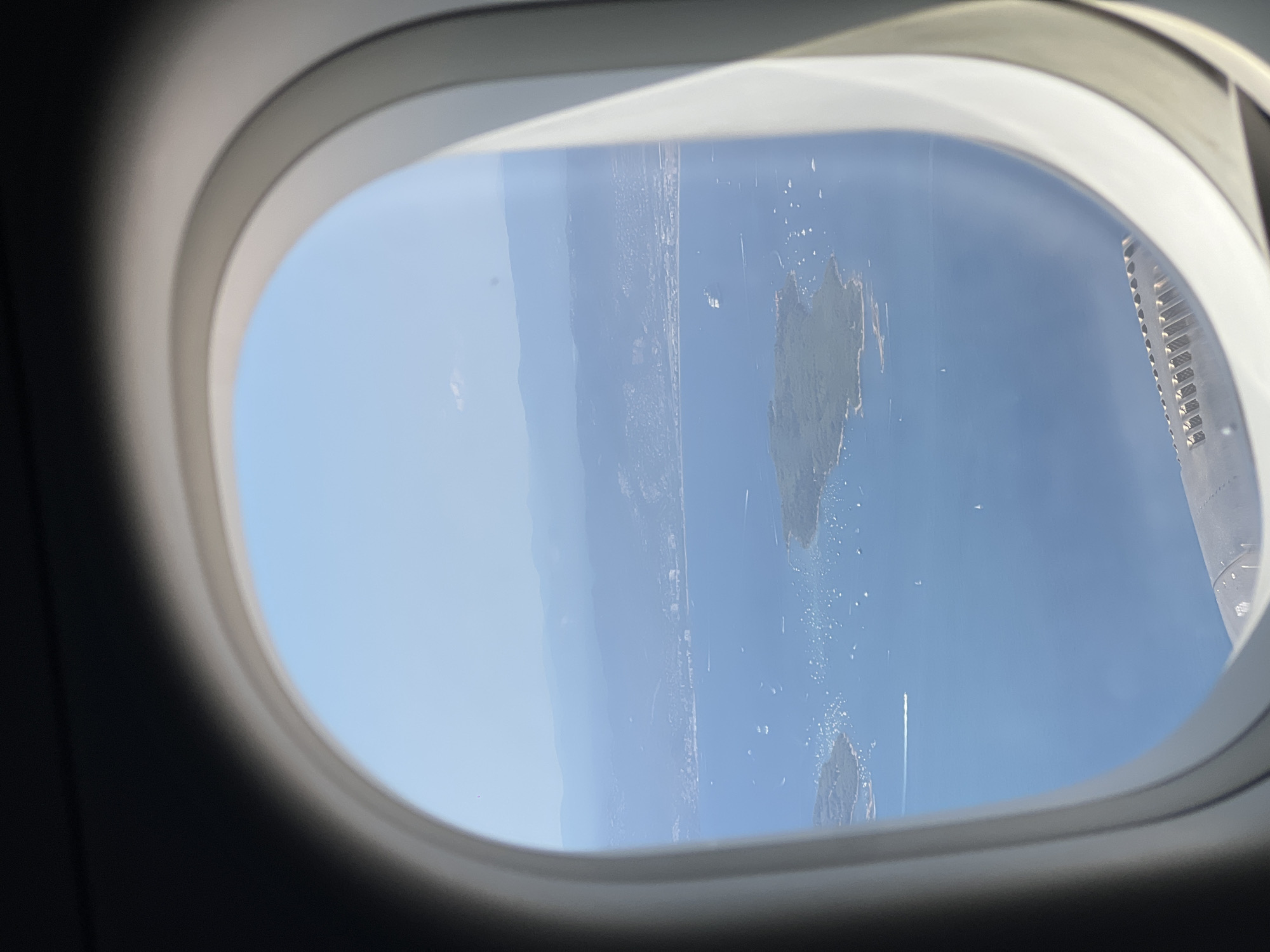 The image is an aerial view from an airplane window looking out over the Mediterranean Sea, towards the coastline of Saint-Laurent-du-Var, France. The foreground is dominated by the rounded edge of the airplane window, with its white interior frame and dark exterior shading.
In the middle ground, the blue expanse of the sea is dotted with numerous white sailboats and a few motorboats. A medium-sized white ferry or cargo ship is visible in the distance on the water. A green, tree-covered island is situated in the lower left of the frame, with more sailboats anchored around its shores.
The background features a hazy, mountainous coastline. The town or city of Saint-Laurent-du-Var is visible along the coast, characterized by a strip of buildings and structures nestled against the hills. The sky above is a clear, pale blue, with a faint, wispy cloud in the upper left quadrant. Part of the airplane's wing, with its textured surface, is visible along the bottom edge of the frame. The overall impression is one of a clear, sunny day with calm seas.