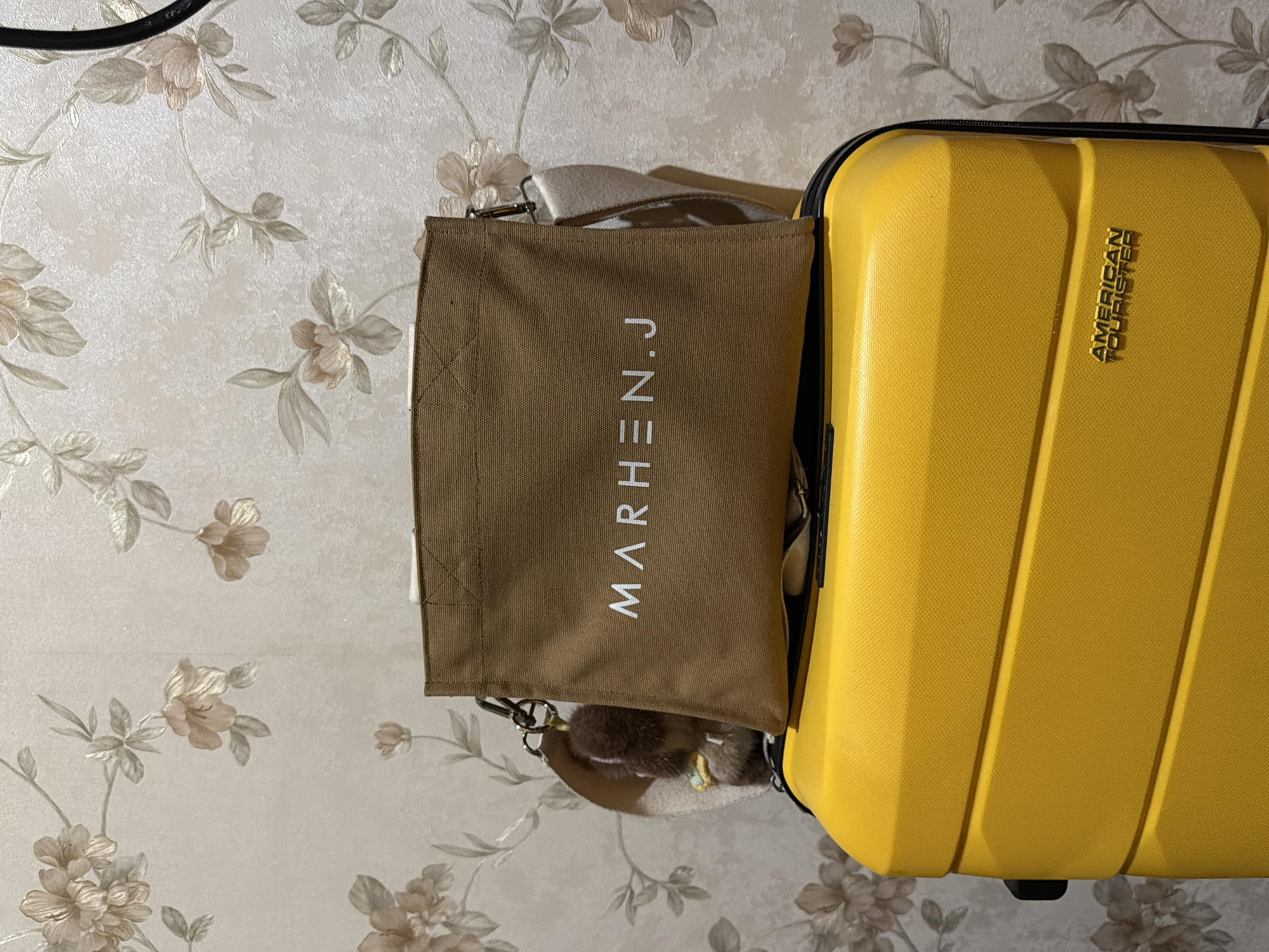 The scene depicts a yellow American Tourister suitcase standing upright. A brown MARHEN.J tote bag hangs from its handle. The suitcase and tote bag rest on a surface with a floral wallpaper background. The location is East Jakarta, Indonesia.