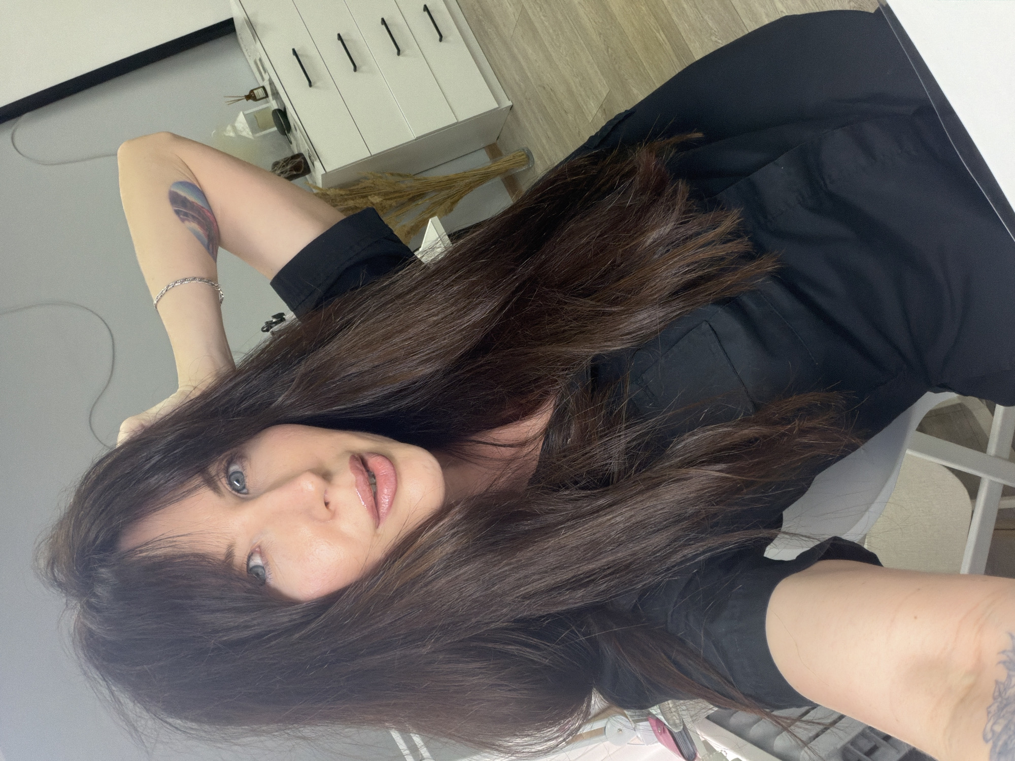 A woman with long brown hair and light blue eyes is posing for a selfie indoors. She is wearing a black shirt or dress. She has a tattoo on her left arm and a silver bracelet on her left wrist. In the background, there is a white dresser with black handles, a vase with dried flowers, and a white chair. The location is likely Perm, Russia, as it is referenced in the prompt.