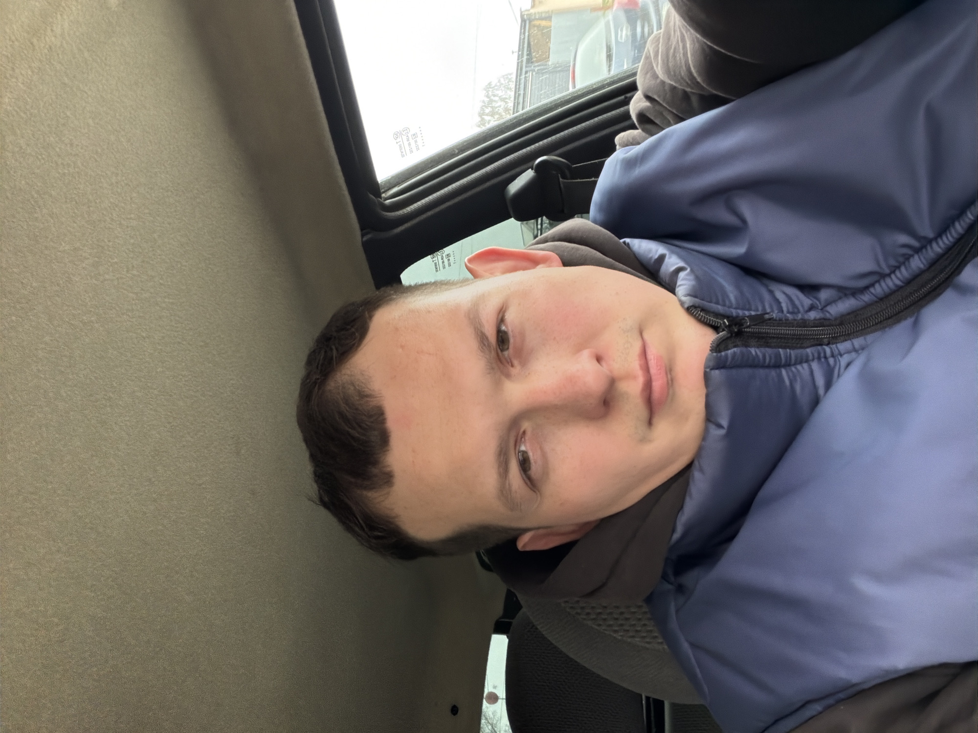 The image is a sideways close-up portrait of a young adult male, appearing to be a selfie taken from inside a vehicle. The man has short, dark hair, fair skin, and brown eyes, looking directly into the camera with a neutral to slightly contemplative expression. He is dressed in a dark blue or slate-grey puffy jacket, worn over a dark, possibly hooded, inner layer.
The setting is the interior of what appears to be a car or similar vehicle. A textured, light-colored headliner or ceiling is visible on the left. To the right, a window reveals an outdoor scene. The window glass bears several stickers with technical markings, including numbers like "23755" and symbols such as "DOT" and "CCC," which are typical of vehicle safety or manufacturing labels. Outside, the weather appears overcast and cold, with visible snow or ice on what looks like a building or structure. Blurred architectural elements are present in the background. Parts of the vehicle's interior, including a dark seat and a seatbelt buckle mechanism, are also discernible. The overall lighting is soft and diffused, consistent with an overcast day.