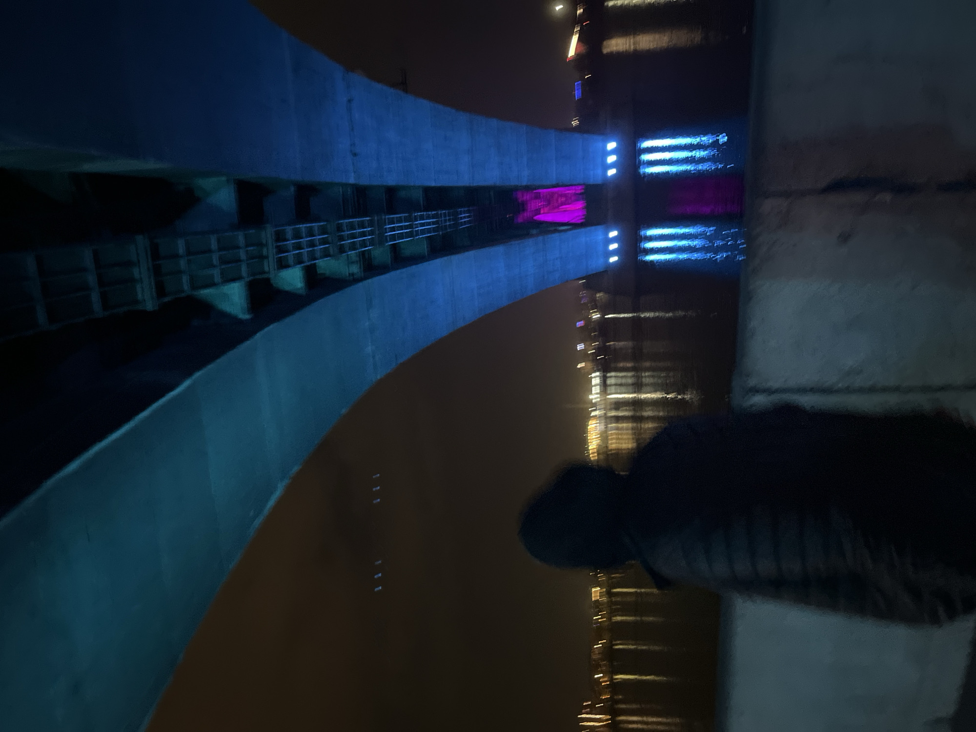The image shows a night view of the Vinogradovsky Bridge in Krasnoyarsk, Russia. The bridge's blue-lit concrete arches and the support structure are visible on the left side of the frame. Lights from buildings and reflections on the Yenisey River are seen in the background. A person is standing on the right side of the image, near a concrete barrier. Their face is obscured, but they appear to be wearing a dark hooded jacket or shirt.