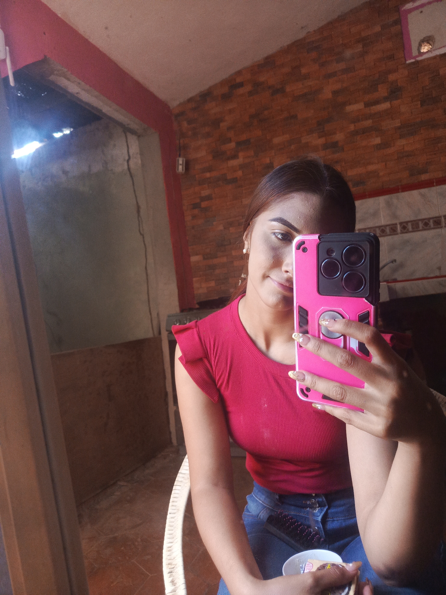 A woman is taking a selfie with a pink-cased smartphone. She is wearing a red, short-sleeved, ruffled top and blue jeans. She is holding a hairbrush and a small cup or container. The setting appears to be indoors, with a brick wall in the background and a doorway to an outdoor area with weathered concrete. The exact location in Juan Griego, Venezuela, cannot be determined from the image.
