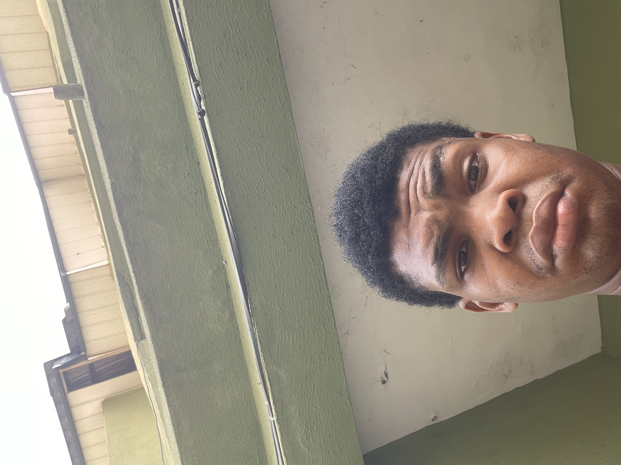 A selfie taken from below of a Black man with short, dark, curly hair. He has dark eyes, a broad nose, and full lips. He is looking directly at the camera with a slightly furrowed brow. The background includes a green building facade with vertical lines and a white ceiling. The location is Ado, Nigeria.