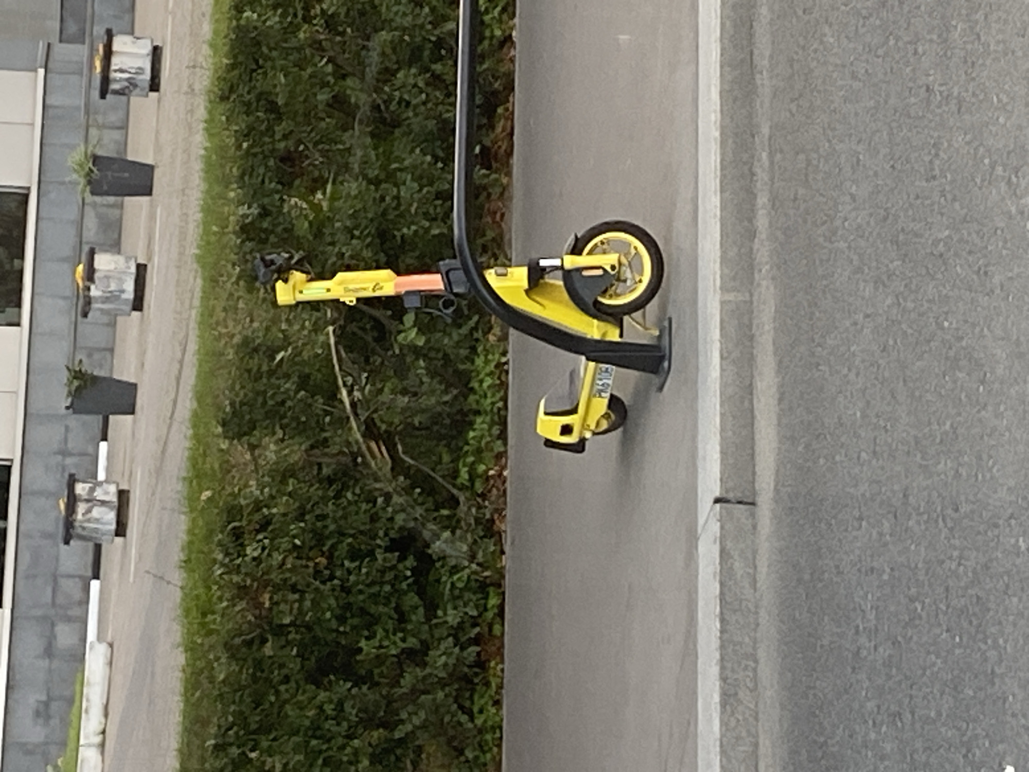 A yellow electric scooter with a black stand is parked on a paved walkway beside a dense green hedge. The scooter has a number plate that appears to read "PK6108". Behind the hedge, a modern building with a grey facade and windows is visible. In front of the building, several large, cylindrical planters are arranged in a line, some containing plants. The walkway is separated from the road by a grey curb. The image appears to have been taken in Moscow, Russia.