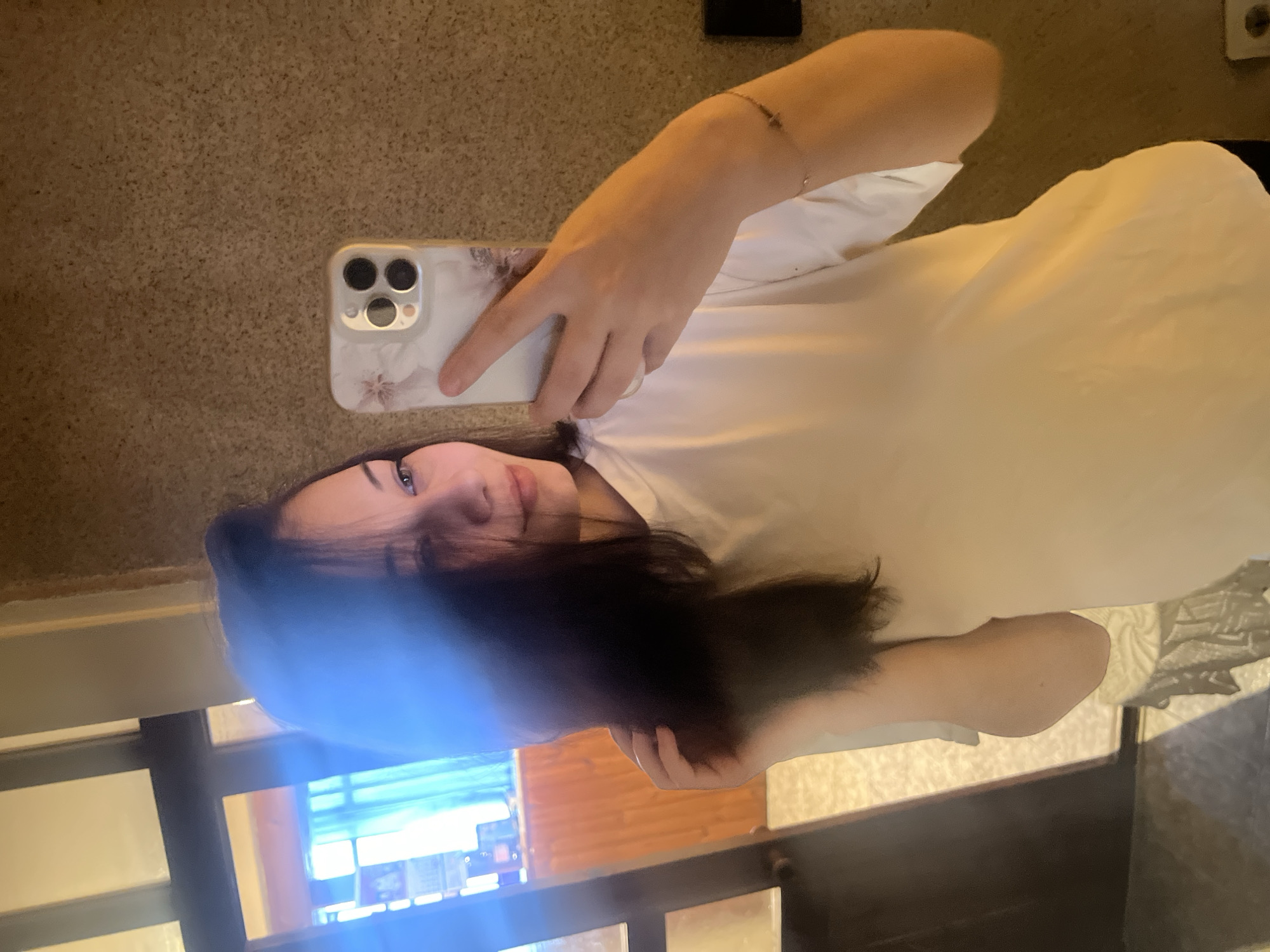 A person is taking a selfie indoors. The person has long dark hair, fair skin, and is wearing a light-colored shirt. The person is holding a cell phone with a floral design on the back. A window with horizontal bars is visible in the background with a building with a blue awning. The objects in the background of the window appear to match the location of Elena, Bulgaria.
