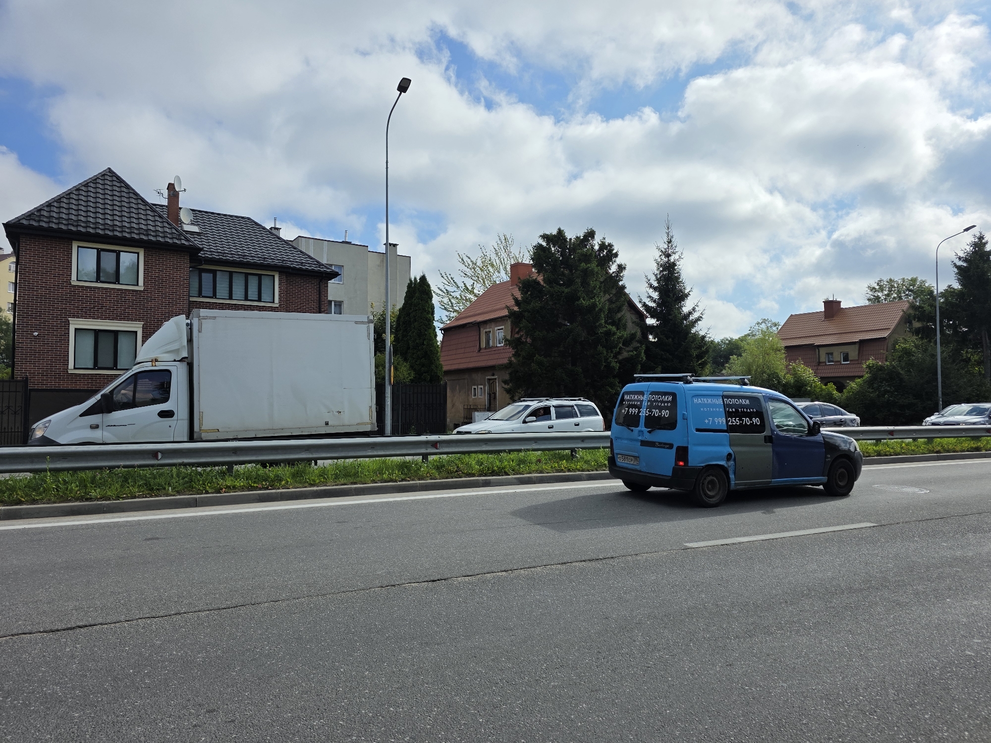 The image depicts a roadside scene in the city of Kaliningrad, Russia, under a cloudy sky. A grey asphalt road with white lane markings occupies the foreground. In the middle ground, a silver guardrail runs along the edge of the road, separating it from a grassy verge. Beyond the guardrail, a white box truck is parked, its rear half visible. To the right of the truck, a blue and grey van is driving away from the camera. The van has text and a phone number printed on its side. In the background, there are several buildings, including a red brick house with a dark tiled roof, and other residential structures. Trees and utility poles are also present in the scene.