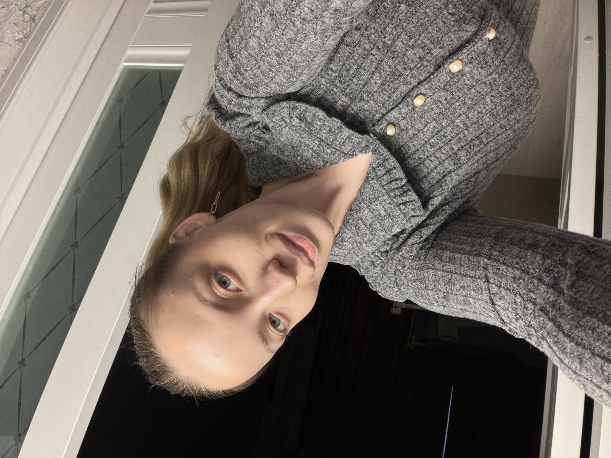 A fair-skinned woman with blonde hair is posing for a selfie. She is wearing a grey sweater with pearl buttons. The background appears to be a window and a dark space. There is no indication of the location being Reshetnikovo, Russia.