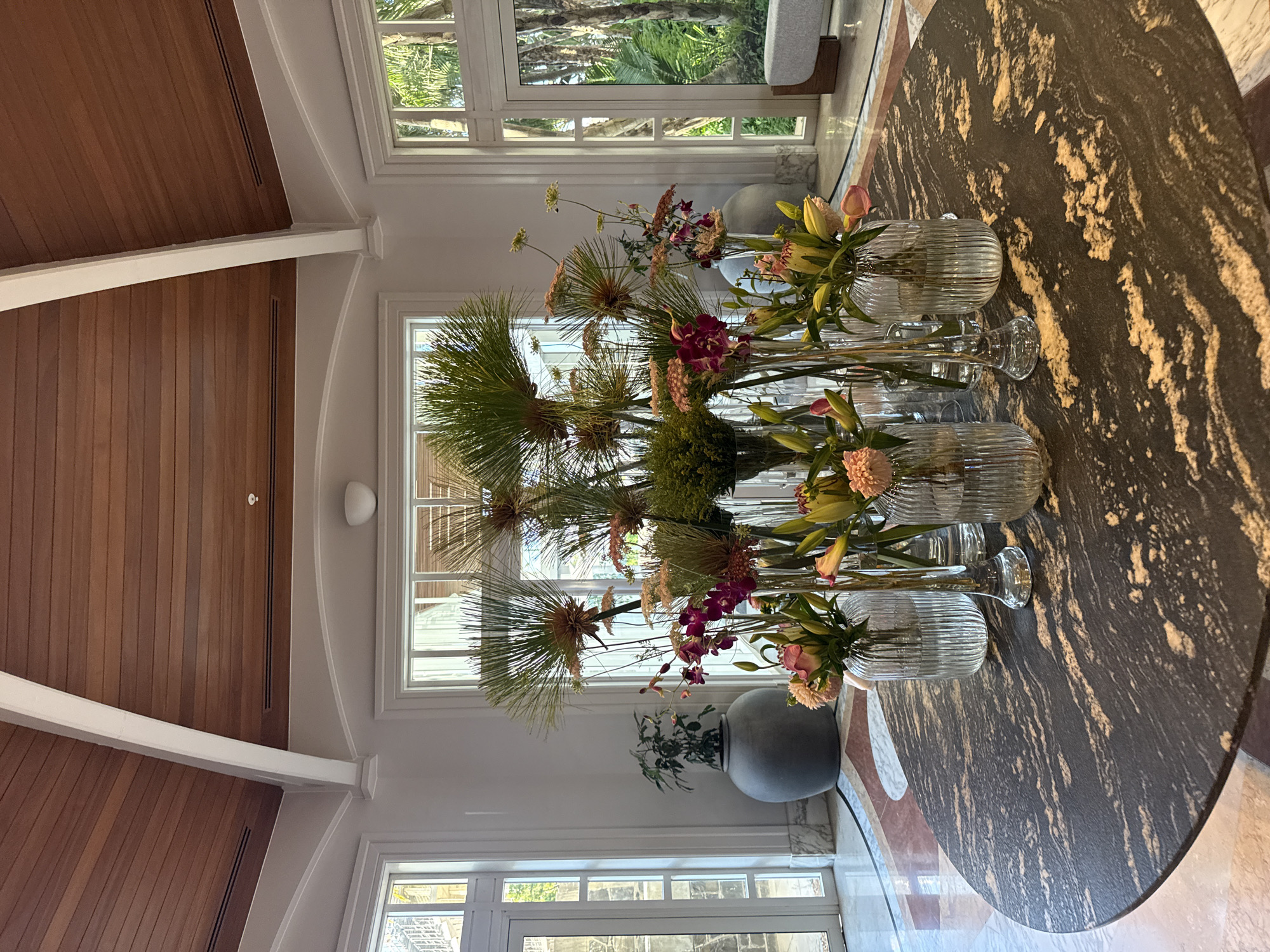 The image depicts an interior space featuring a large round table with a dark, granite-like surface adorned with multiple glass vases filled with floral arrangements. The flowers consist of assorted blooms and greenery, including what appears to be protea. Behind the table is a series of windows offering a view of lush greenery, potentially palm trees. The architecture of the building features white trim and a wood-paneled ceiling. The scene suggests a well-lit, possibly upscale interior setting, and the presence of the protea flowers points towards a location in or around Cape Town, South Africa, given their indigenous origin. The large, gray potted plant in the background adds to the interior's aesthetic. The composition is captured from a low angle, slightly rotated. There are no people visible in the frame.