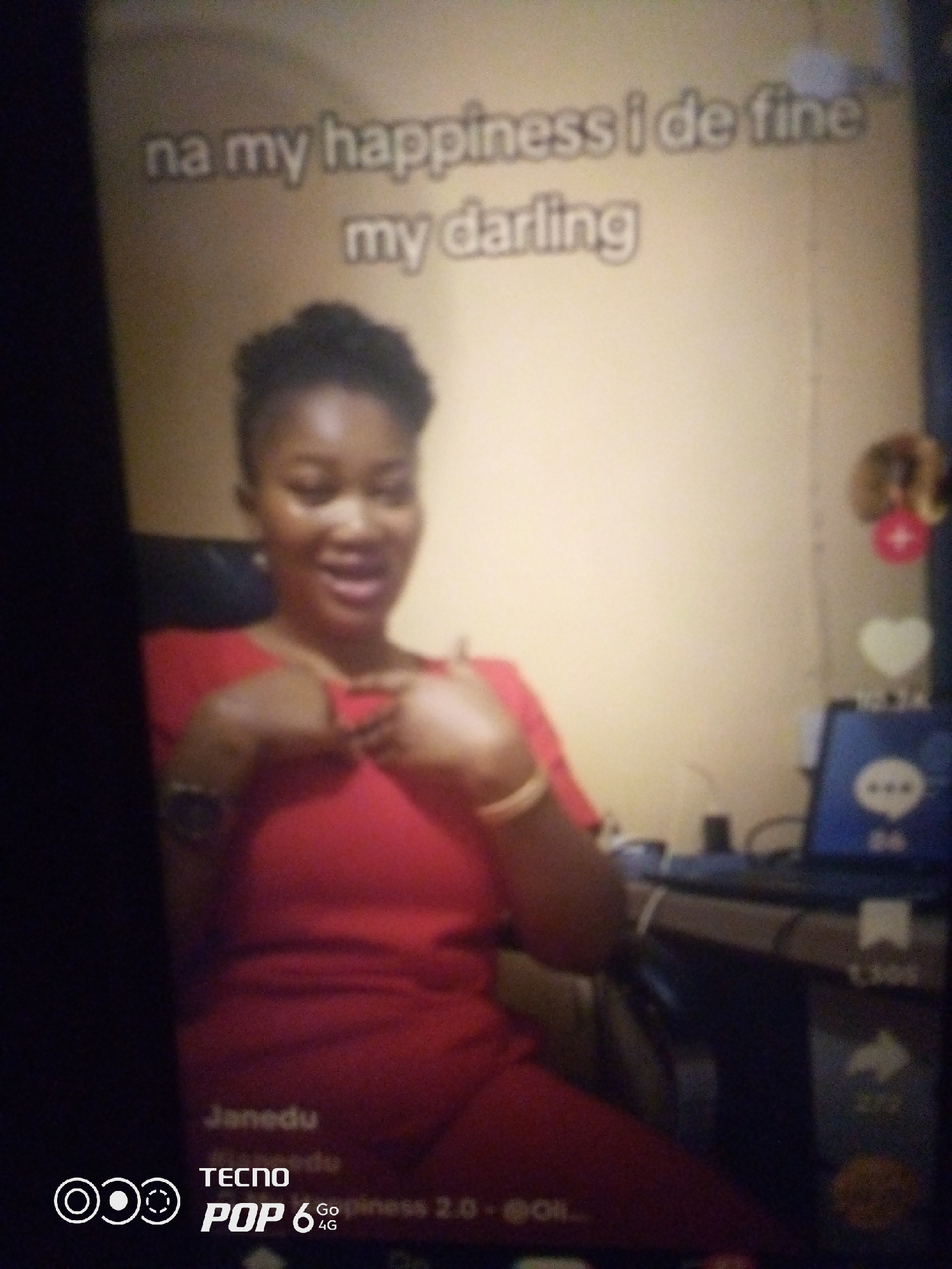 The image shows a woman in a red outfit, with her hands on her chest. The background is a light-colored wall with some text that is difficult to read due to the blurriness of the image. There is a laptop visible on a desk to the right of the woman, displaying a blue screen with some icons. The woman appears to be smiling. The image also contains a watermark at the bottom left with the text "TECNO POP 6 Go 4G". The text at the top of the image reads "na my happiness i de fine my darling". There are no visible elements in the image that would indicate the location in the city of Howayi, Nigeria.