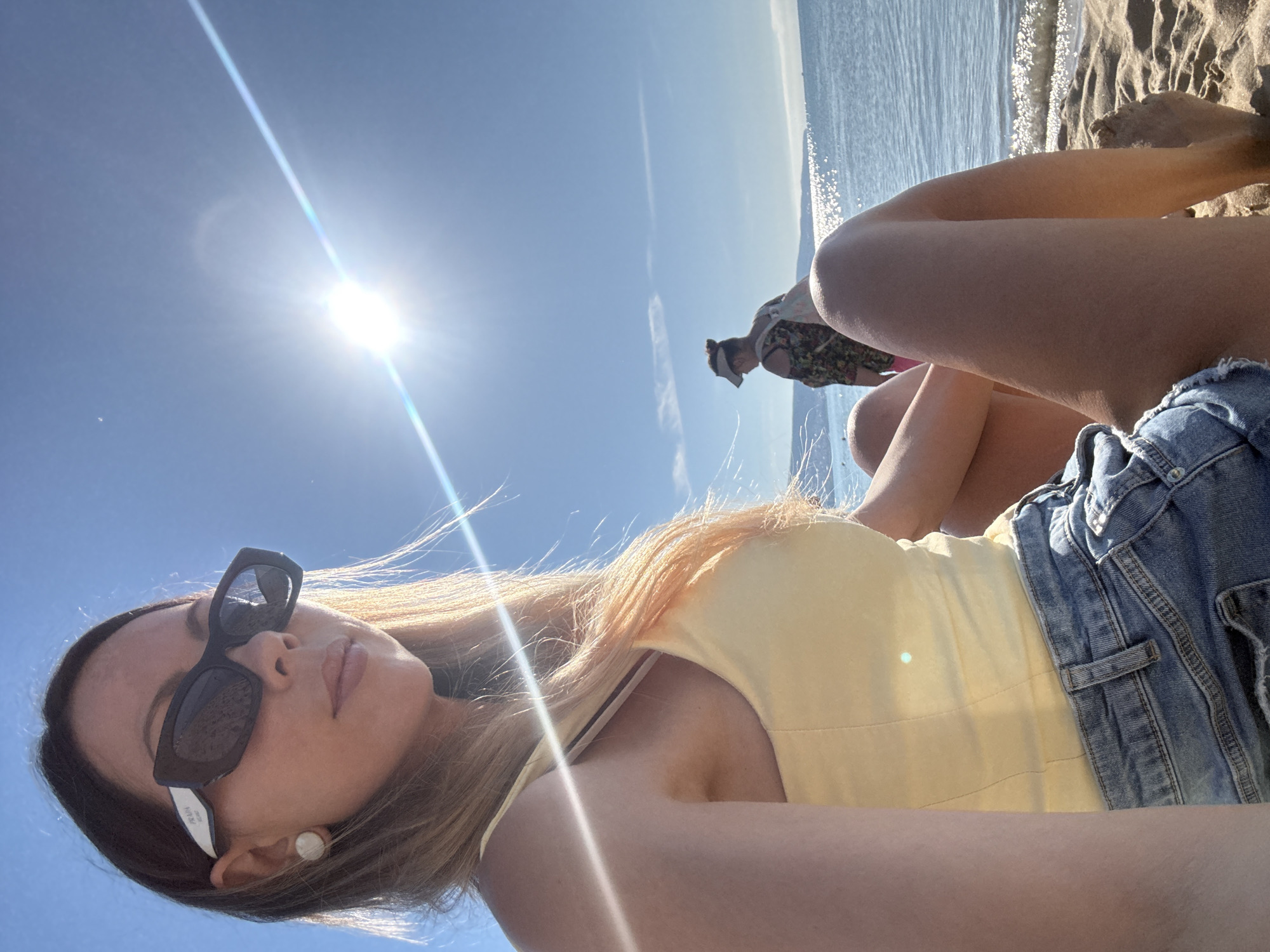 A woman is sitting on a sandy beach, facing forward with her legs crossed. She is wearing a yellow top and denim shorts. The sun is shining brightly in the blue sky, casting a glare on the water and the surrounding environment. In the background, another person is visible near the water. The location appears to be the coast of Nesebar, Bulgaria, a historic city known for its beaches and ancient architecture.