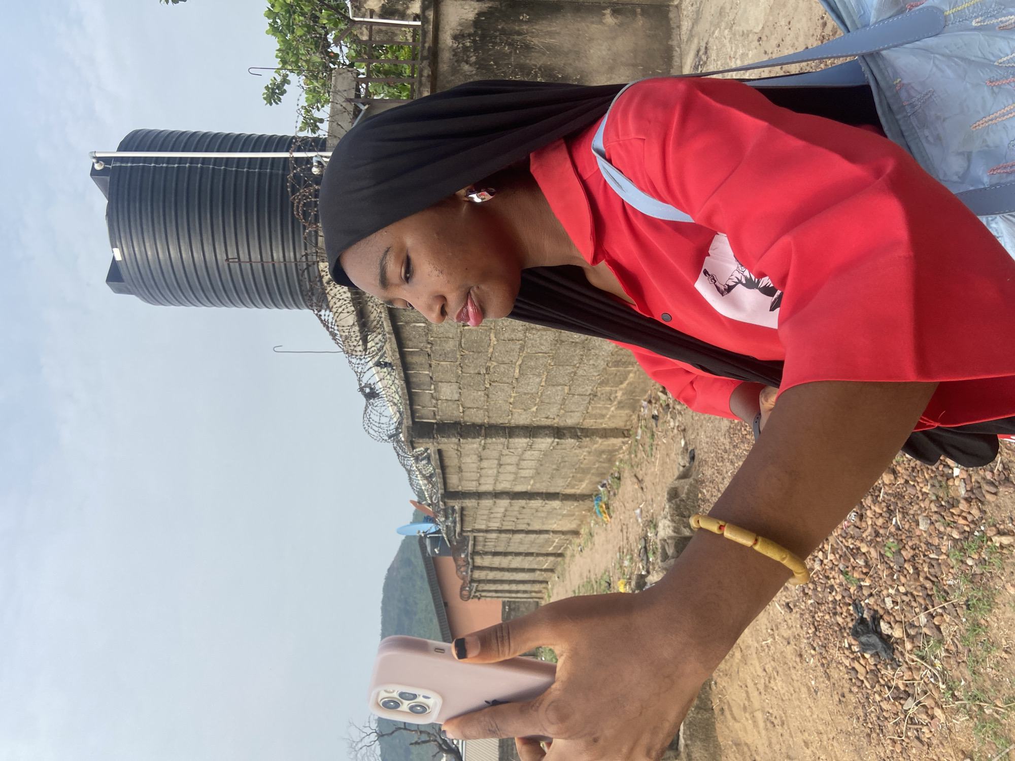 The image features a young person with dark skin, positioned outdoors, focused on a light pink smartphone held in their right hand. They are wearing a black head covering that drapes over their shoulders, and a vibrant red long-sleeved shirt. A white graphic, possibly depicting a figure with a rifle, is partially visible on their shirt. A light blue strap from a bag hangs over their left shoulder, and they accessorize with silver earrings and a light-colored, beaded bracelet on their right wrist. Their lips are slightly parted, indicating concentration.
In the mid-ground, a sturdy concrete block fence topped with coils of razor wire marks a boundary. Beyond the fence, the ground is unpaved and rocky, with glimpses of a building featuring an orange roof and a satellite dish. A prominent black, ribbed cylindrical water tank is visible to the left, standing tall against the sky. The distant background reveals a range of green, tree-covered hills under a clear, pale blue sky, suggesting a bright daytime setting. The foreground consists of dirt and scattered gravel. The overall scene reflects an outdoor environment, possibly in a semi-urban or rural area like Lokoja, Nigeria. No readable text is visible on any surfaces.