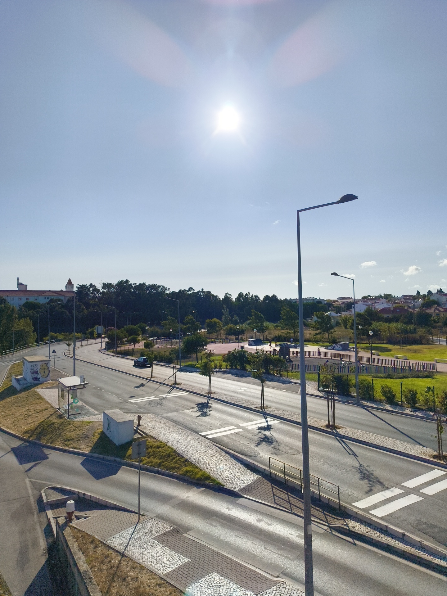 The image captures an urban landscape bathed in bright sunlight, likely taken from an elevated vantage point. The sky is a clear, bright blue with the sun prominently visible, creating lens flares. In the foreground, a paved road with distinct white crosswalk markings curves through the scene. Several streetlights stand tall along the roadside. To the left of the road, there's a bus stop shelter with a small kiosk next to it, and further back, a building with a red-tiled roof and pale walls is visible.
The middle ground features a park area with green grass, trees, and recreational facilities, including what appears to be a skate park with ramps and benches. A dark-colored car is parked on the road. The background consists of a dense line of trees and residential buildings with red roofs, characteristic of Portuguese architecture. The environment appears to be a well-maintained urban area with a mix of residential and recreational spaces.
Based on the architectural style, particularly the red-tiled roofs, and the presence of a bus stop, this scene is likely located in the city of Cascais, Portugal.