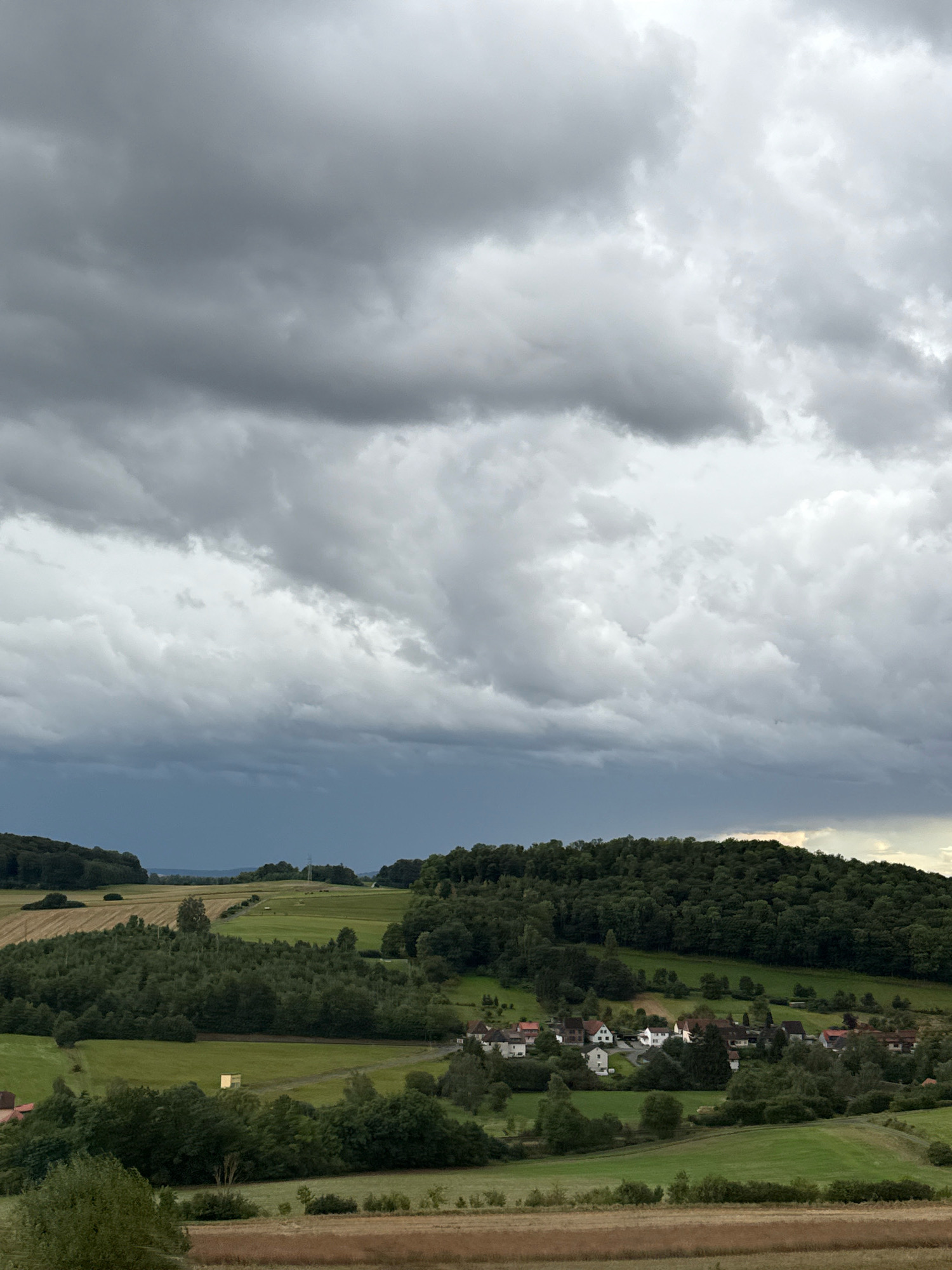 The image depicts a rural landscape under a dramatic, overcast sky. In the foreground, there are fields of dry grass and some trees. The middle ground features rolling green hills dotted with trees and a small village with several houses. A dense forest covers the top of a hill on the right side of the image. The sky is filled with dark, heavy clouds, suggesting an impending storm. The overall impression is one of a serene but somewhat foreboding natural scene. This scene appears to be located in or around the city of Bad Lauterberg im Harz, Germany, a region known for its natural beauty and hilly terrain.
