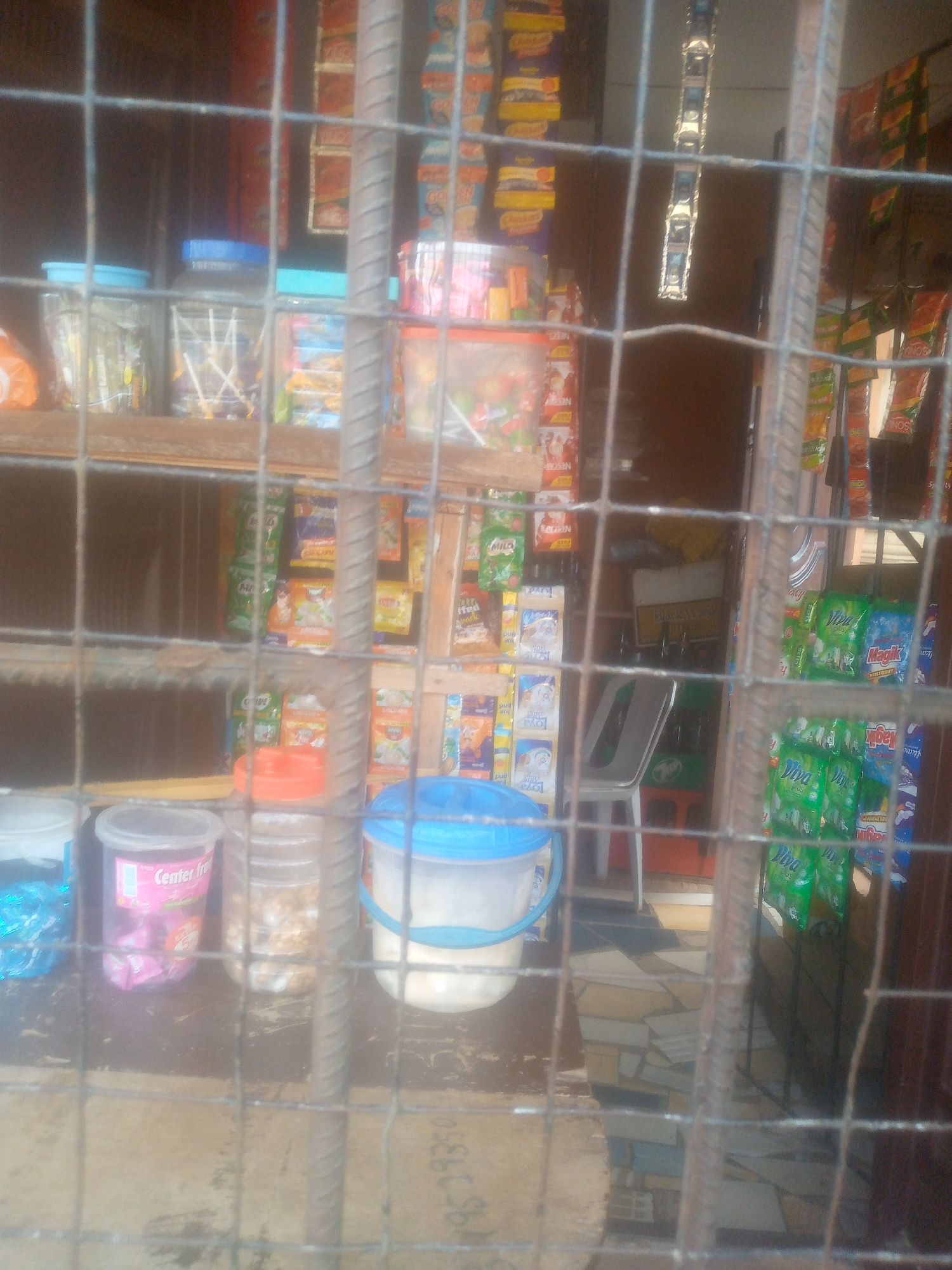 The image depicts the interior of what appears to be a small convenience shop or market stall, viewed through a sturdy metal security grate. The setting is likely an urban or semi-urban area during daylight hours, given the ambient light, though no direct time or weather cues are visible.
The scene is cluttered with various goods displayed on shelves and on a counter. On the top shelf, several clear plastic containers with blue lids hold lollipops or other small candies. Below, a diverse assortment of packaged products is visible, including sachets and small cartons of food and beverages. On the counter in the foreground, there are several plastic containers with various items inside, including one pink container labeled "Center fro" (likely "Center fresh" or "Center fruit") and another large white bucket with a blue handle and lid, containing a white substance. To the right, green and blue packaged items, possibly detergents or household cleaning products, are prominently displayed, with brands such as "Viva PLUS" and "Magik SUPER BRIGHT" being discernable. Other visible brand names include "MILO," "Loya," and "NESCAFÉ."
In the background, a white plastic chair and a dark crate can be seen, suggesting a seating area or storage. The metal grate heavily obscures the view, making many details blurry and indistinct. No people are present, and no specific activity is taking place; the image captures a static display of merchandise. A number, "9519350," is partially visible on a surface in the lower center of the image.