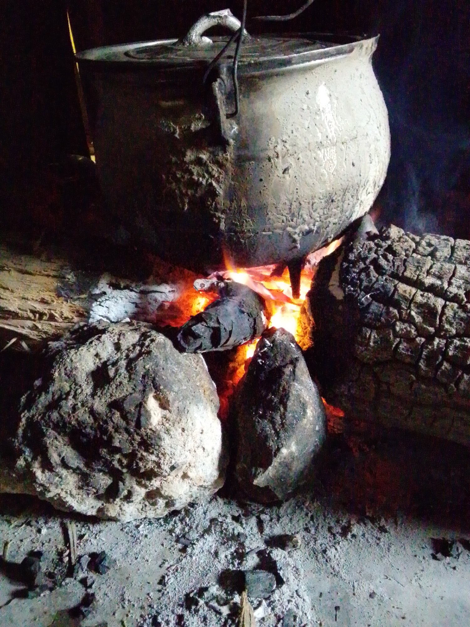 The image depicts a traditional outdoor cooking scene, likely in Jalingo, Nigeria, where a large, weathered pot is being heated over an open fire. The pot, made of a dark, possibly metal material, is encrusted with soot and residue, indicating heavy use. It is suspended above burning logs and glowing embers, with flames visible beneath. Several large, irregular stones surround the fire pit, serving as supports for the pot or as part of the cooking setup. Two substantial pieces of charred wood, likely logs, are positioned next to the fire, one showing the cracked and blackened surface of burnt timber. The ground is covered in ash and debris, typical of an outdoor fire. The lighting suggests it is either dusk or nighttime, with the fire providing the primary illumination. There are no people visible in the image, focusing solely on the cooking apparatus and its immediate surroundings.