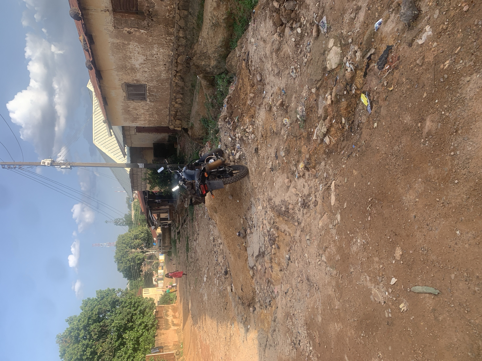 This media file depicts a scene in Jos, Nigeria. The orientation of the image is portrait, with the perspective from eye level, suggesting it was captured by someone standing. The scene is primarily an outdoor environment, characterized by a dirt road and a rough, unpaved terrain with loose soil, rocks, and scattered debris, indicative of an undeveloped area.
In the background, to the left, there is a weathered, two-story building with peeling paint and a metal roof. It appears to be an older structure, possibly residential or commercial. Adjacent to it, another building with a corrugated metal roof is visible. Further in the distance, telephone poles with wires stretch across the sky, and communication towers are visible, suggesting some level of infrastructure. Lush green trees are scattered throughout the scene, particularly on the left side, adding a touch of nature to the otherwise rugged landscape.
A black motorcycle is parked on the dirt road, serving as a focal point. Its headlights are on, suggesting it might be daytime, possibly late afternoon given the shadows. A person in a red garment is walking away from the motorcycle on the road, and another person is visible in the distance near some greenery. The presence of these individuals implies everyday life and activity in this location.
The sky is partly cloudy with some bright blue patches, indicating fair weather. The overall impression is one of a rural or semi-urban area undergoing development or experiencing less infrastructure. There is no visible text in the image. The scattered debris and the state of the buildings and road might suggest a lack of consistent maintenance or waste management in the area. The scene appears to be from a warm, possibly sunny day.