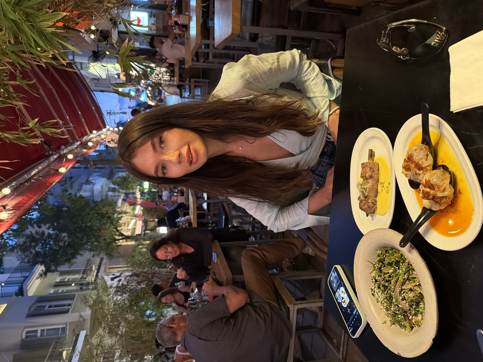 The image captures a woman with long, dark hair sitting at a table in an outdoor restaurant in Tel Aviv, Israel. She is wearing a light blue wrap top and denim shorts. In front of her, on the black table, are several plates of food: a salad in a large bowl, a piece of meat on a skewer with a yellow sauce, and two dumplings in a sauce. A smartphone is also visible on the table, displaying images of food. In the background, other patrons are seated at tables, and the restaurant appears to be on a street with buildings and trees. The lighting suggests it is evening or dusk.
