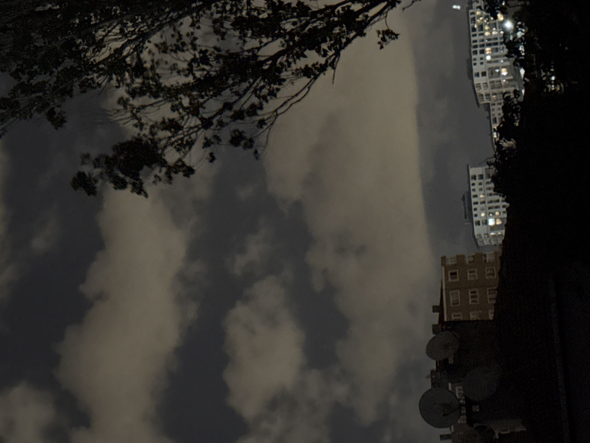 This is a nighttime, low-light photograph of the city of Gungoren, Turkey. The foreground is dominated by the dark silhouettes of trees and the roof of a building adorned with multiple satellite dishes. In the midground, several multi-story apartment buildings are visible, their windows illuminated, suggesting occupancy. The background is filled with a cloudy night sky, with patches of lighter clouds contrasting against darker areas. The overall impression is of a residential urban environment at night.