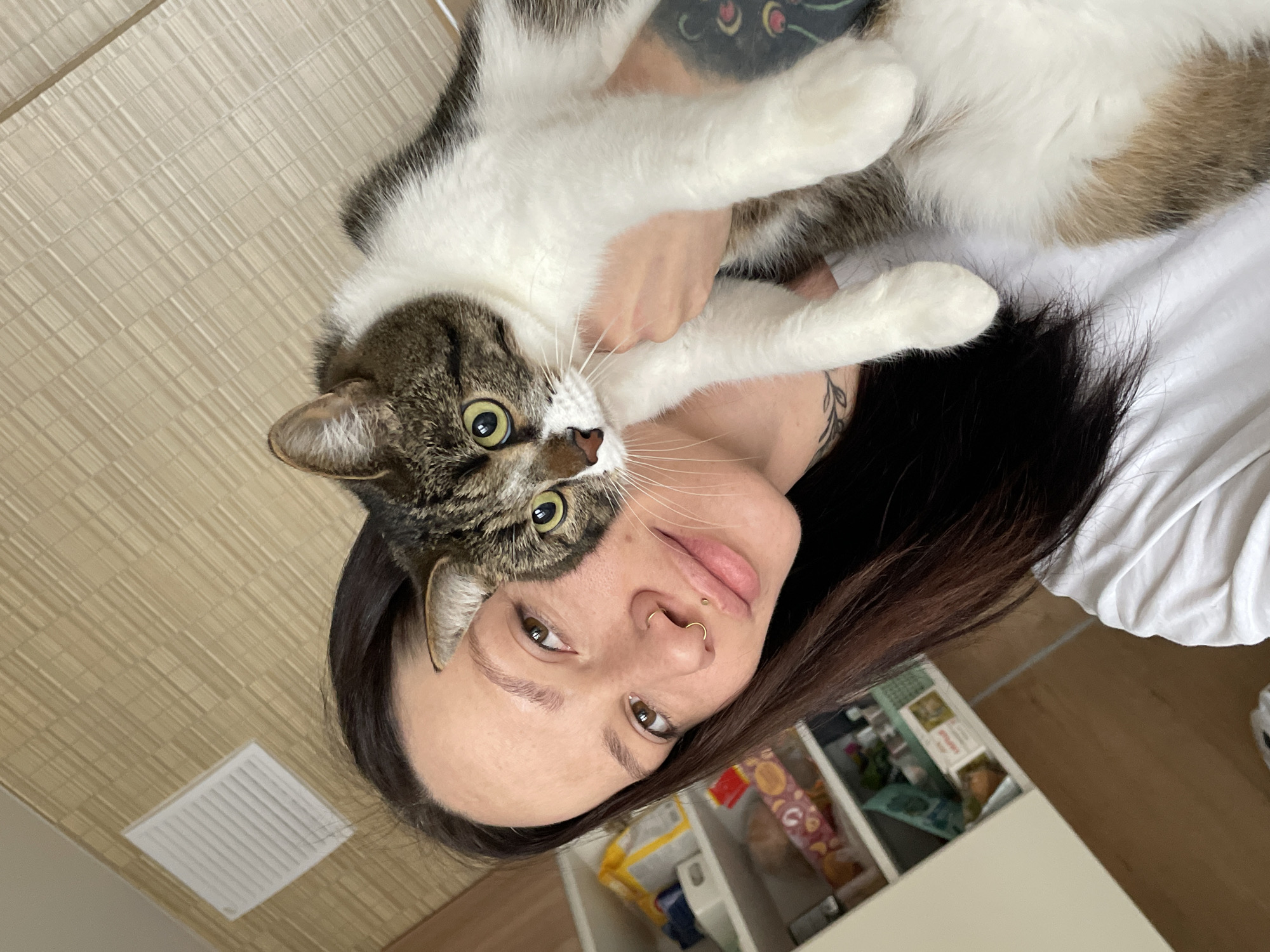 A person with dark hair is holding a cat. The cat is white with brown and black markings. The person has a nose ring and tattoos on their arm and neck. The background includes a light-colored wall, a white cabinet, and a vent on the ceiling. The location is indeterminate.