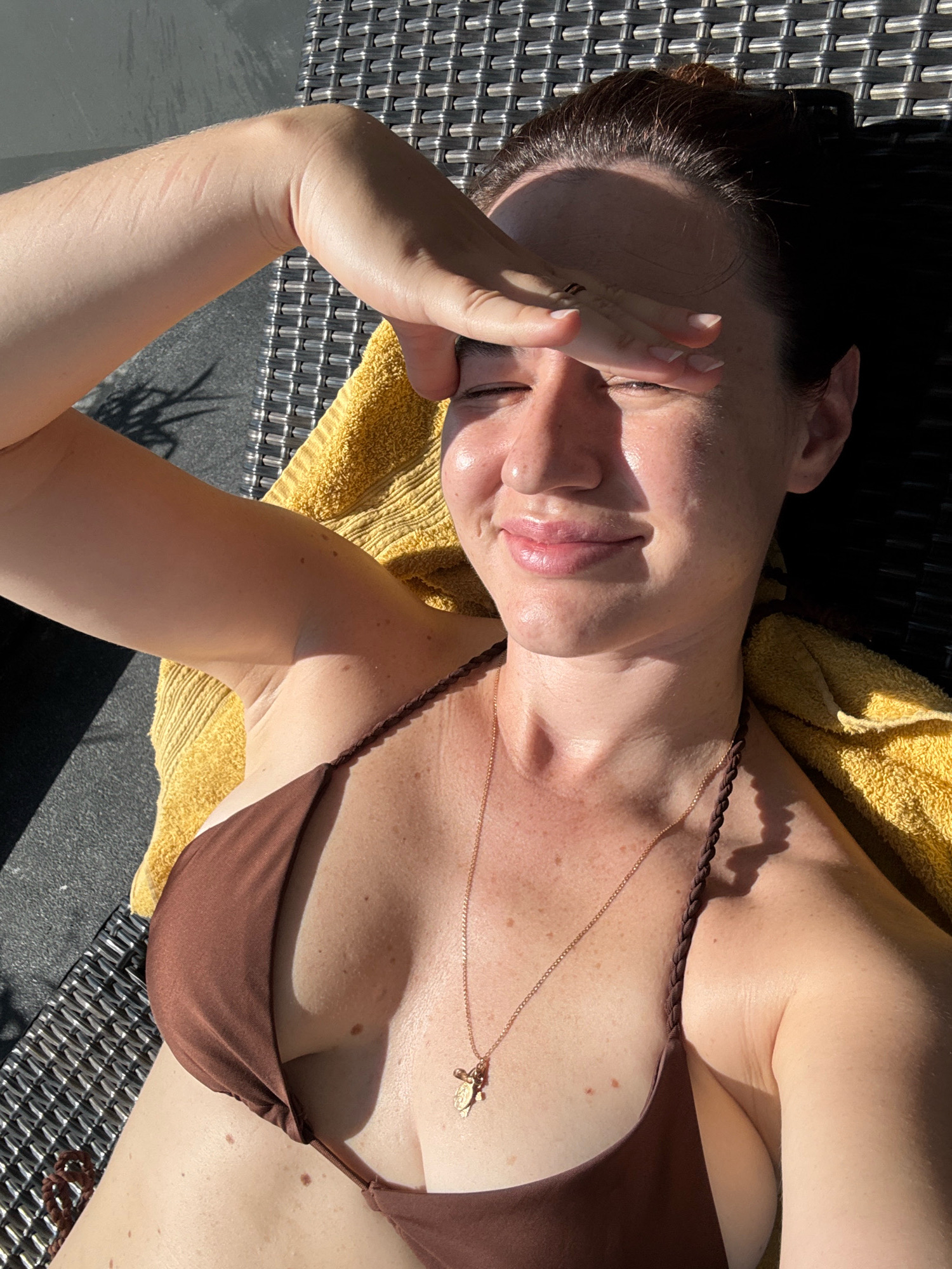 A woman, lying on a lounger covered with a yellow towel, shields her eyes from the sun with her right hand. She is wearing a brown bikini. The background shows a textured surface, likely a patio or deck, with shadows cast by sunlight. The image was taken in Samrong Nuea Subdistrict Municipality, Thailand.
