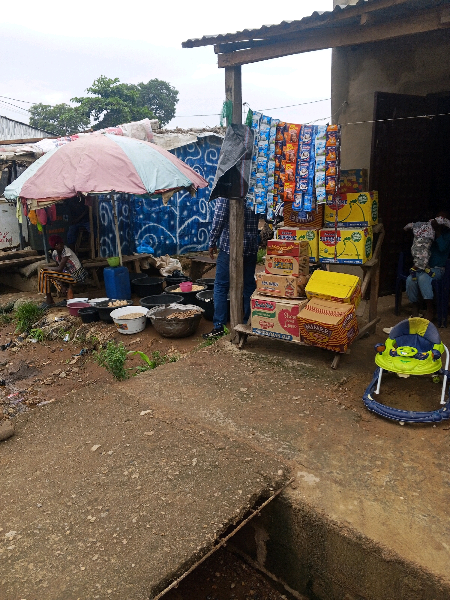 A street market scene is depicted in Lokoja, Nigeria, on a cloudy day. A woman wearing a striped top and pants sits under a large, pink and light blue umbrella. In front of her, on the ground, are several large, dark basins containing various food items such as grains, eggs, and what appears to be dried fish. To the right of the woman, a display of packaged food products, including boxes of noodles and biscuits, is arranged on a wooden shelf. Among these products are several yellow boxes labeled "SUPREME." A green and blue baby walker is placed on the ground in the foreground, to the right of the food display. In the background, the scene transitions to a building with a corrugated iron roof. Several people are visible in the background, some appearing to be customers or vendors, though their actions are not clearly discernible. The ground is unpaved and appears to be a mix of dirt and concrete, with some debris scattered around.