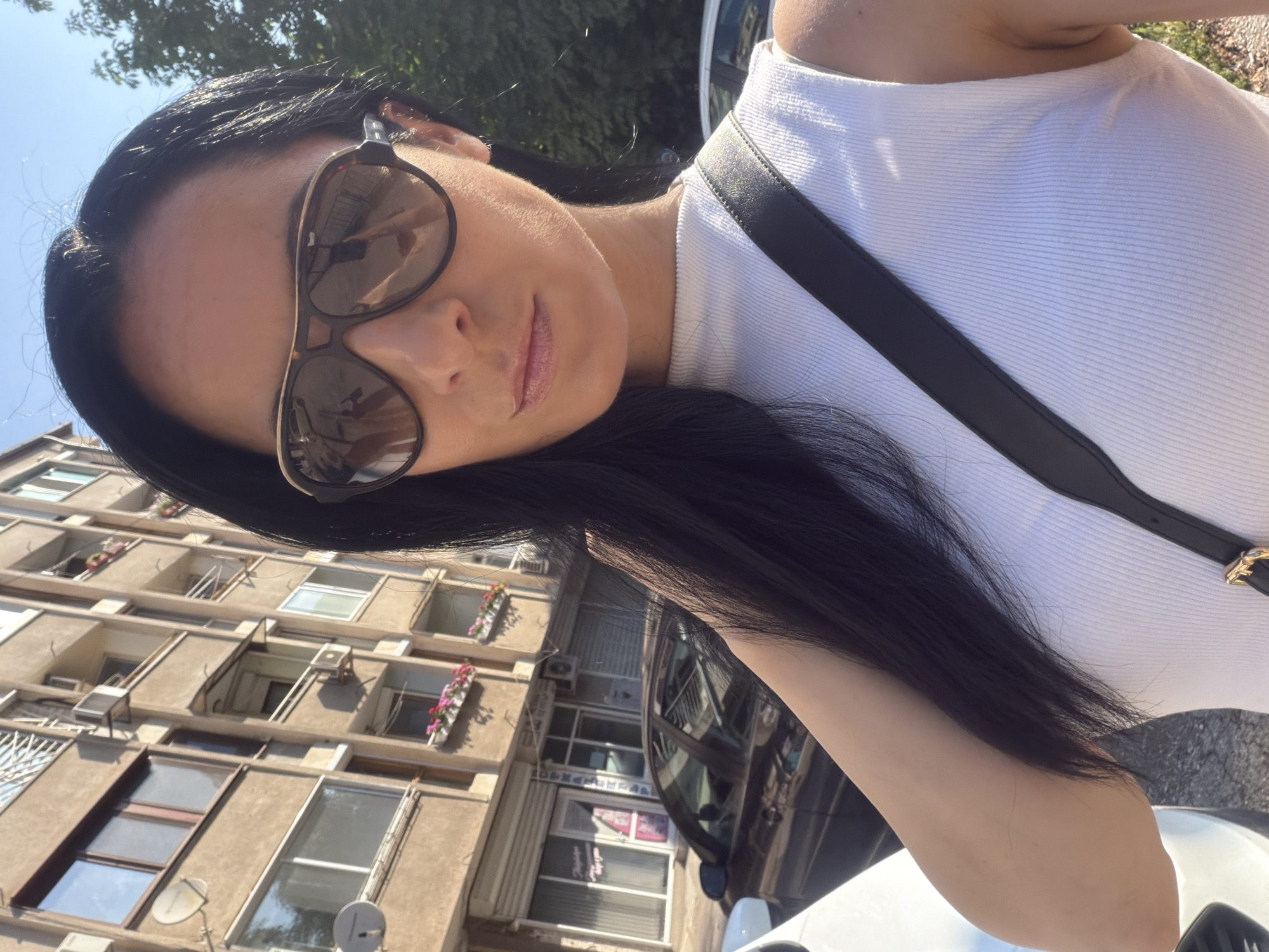 An outdoor selfie captures a person, likely female, with dark hair cascading around her face. She wears large, tortoise-shell sunglasses, which reflect the blue sky and a multi-story building. Her lips are adorned with light-colored gloss. She is dressed in a white, ribbed sleeveless top, with a black strap, possibly from a shoulder bag, crossing her chest.
The background features a concrete-colored building with numerous windows and balconies. Several balconies are decorated with flower boxes containing pink flowers. Satellite dishes are visible on the building's exterior. In the upper left, green tree foliage is seen against a bright, clear blue sky. A white car, possibly reflecting parts of the urban environment, is partially visible at the bottom right. The image is taken in bright daylight, suggesting sunny weather. The location is an urban setting in Pleven, Bulgaria. No readable text is visible in the image.