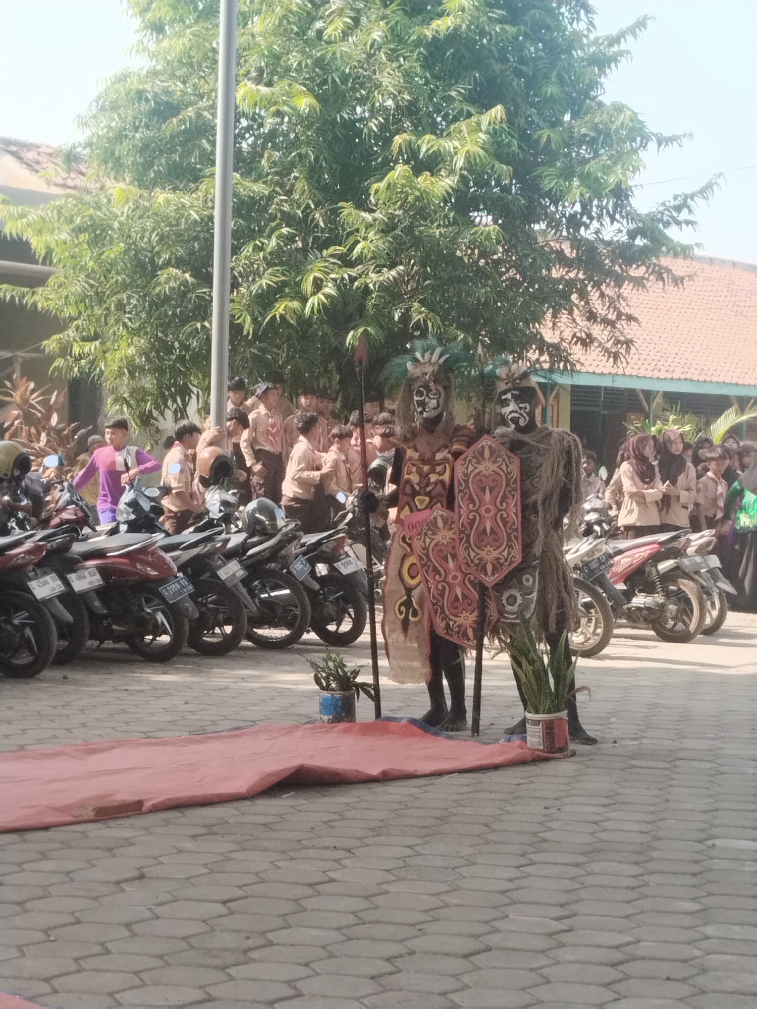 In a courtyard setting, likely at a school in Pakulaut, Indonesia, two individuals dressed in elaborate traditional costumes stand prominently. Their faces are painted with white and black markings, and they wear headdresses adorned with feathers. The individual on the left is draped in a patterned fabric with intricate designs, holding a shield. The individual on the right wears a costume made of what appears to be dried grass or fibers, also holding a shield.
Behind them, a group of students, many in uniform khaki shirts and dark pants or skirts, are gathered, observing the costumed figures. Some students are wearing scout uniforms. A line of parked motorcycles, common for transportation in Indonesian cities, stretches across the background. A red tarp is laid out on the hexagonal patterned pavement in the foreground, suggesting a performance or event area. Trees with lush green foliage provide a natural backdrop, and a building with a terracotta tiled roof is visible in the upper right corner. The overall atmosphere appears to be that of a cultural event or school activity.