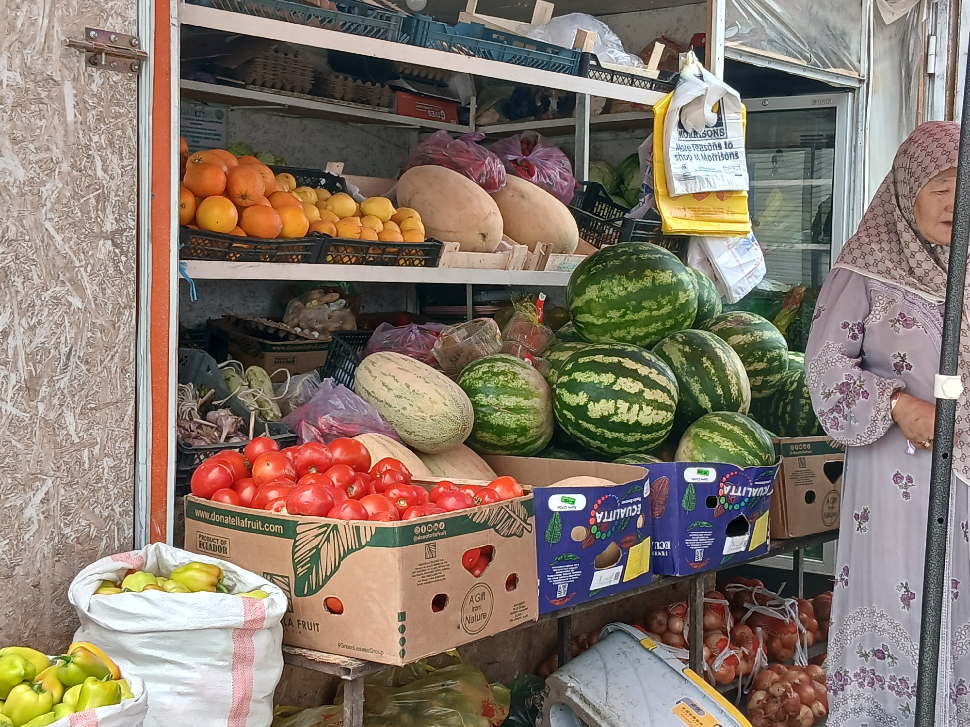 This is a mid-range shot of an outdoor fruit and vegetable stall in Talas City, Kyrgyzstan. The stall appears to be set up against a weathered wooden structure. In the foreground, a large white sack overflows with green bell peppers. To the right of the sack is a cardboard box filled with ripe red tomatoes. Behind the tomatoes, several large watermelons are stacked. A woman wearing a hijab and a patterned dress stands to the right of the frame, partially obscuring the stall. The stall itself is filled with various produce, including oranges, lemons, more watermelons, and what appear to be large melons. Shelving units within the stall hold more produce and some bags. The overall impression is of a bustling market scene.