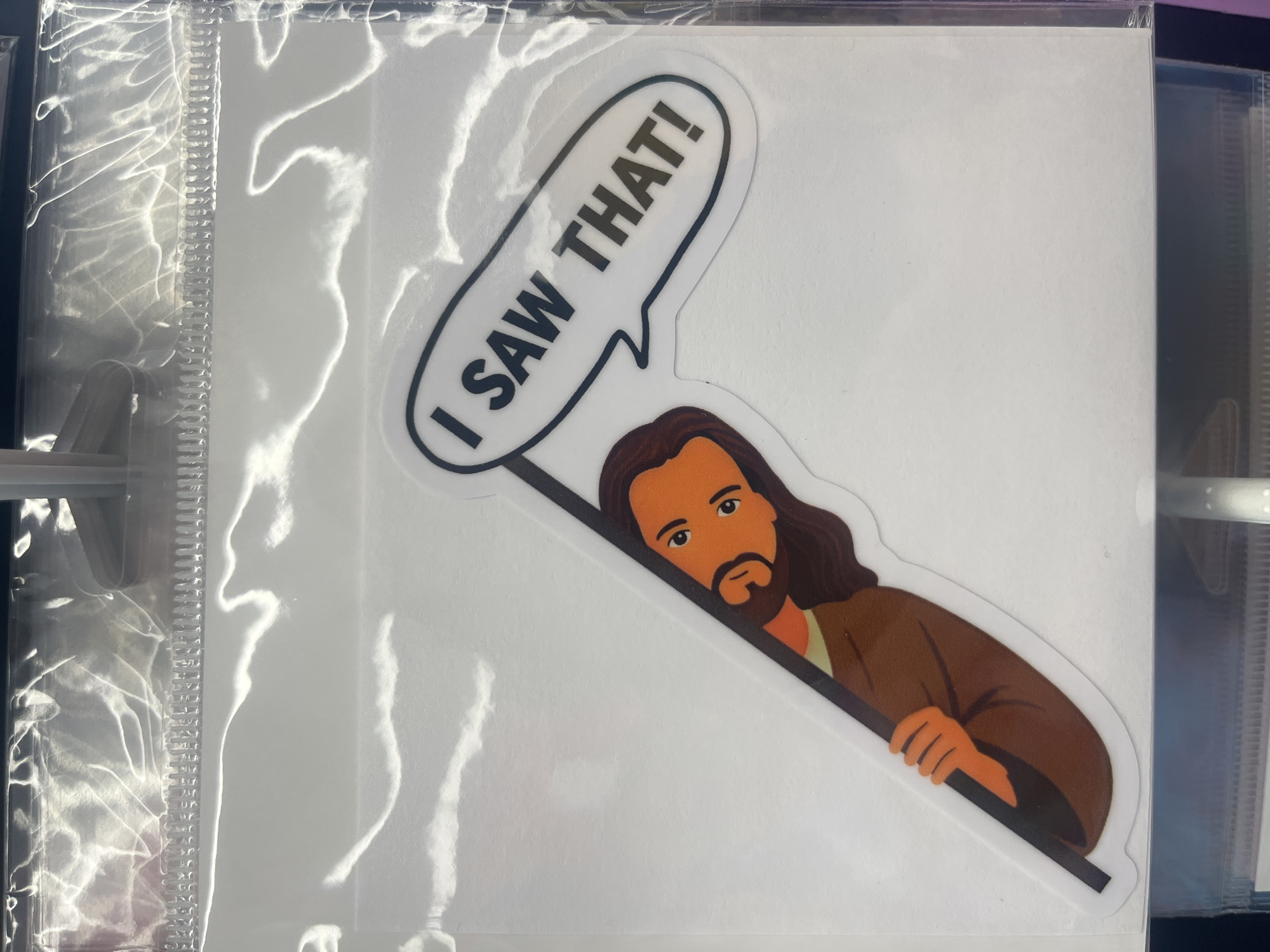 A sticker depicting a cartoonish image of Jesus is shown. The sticker has a white background and a black outline. Jesus is peeking out from behind a black diagonal line, with his face and arm visible. He has brown hair, a beard, and is wearing a brown robe. A speech bubble above his head says "I SAW THAT!" in black capital letters. The sticker is inside a clear plastic package, with a silver zipper-like closure at the top. The sticker is positioned on a white surface, possibly a shelf or display. The image is taken from a slightly high angle, with the plastic packaging reflecting light. There is no identifiable location in Bethany Beach, United States.