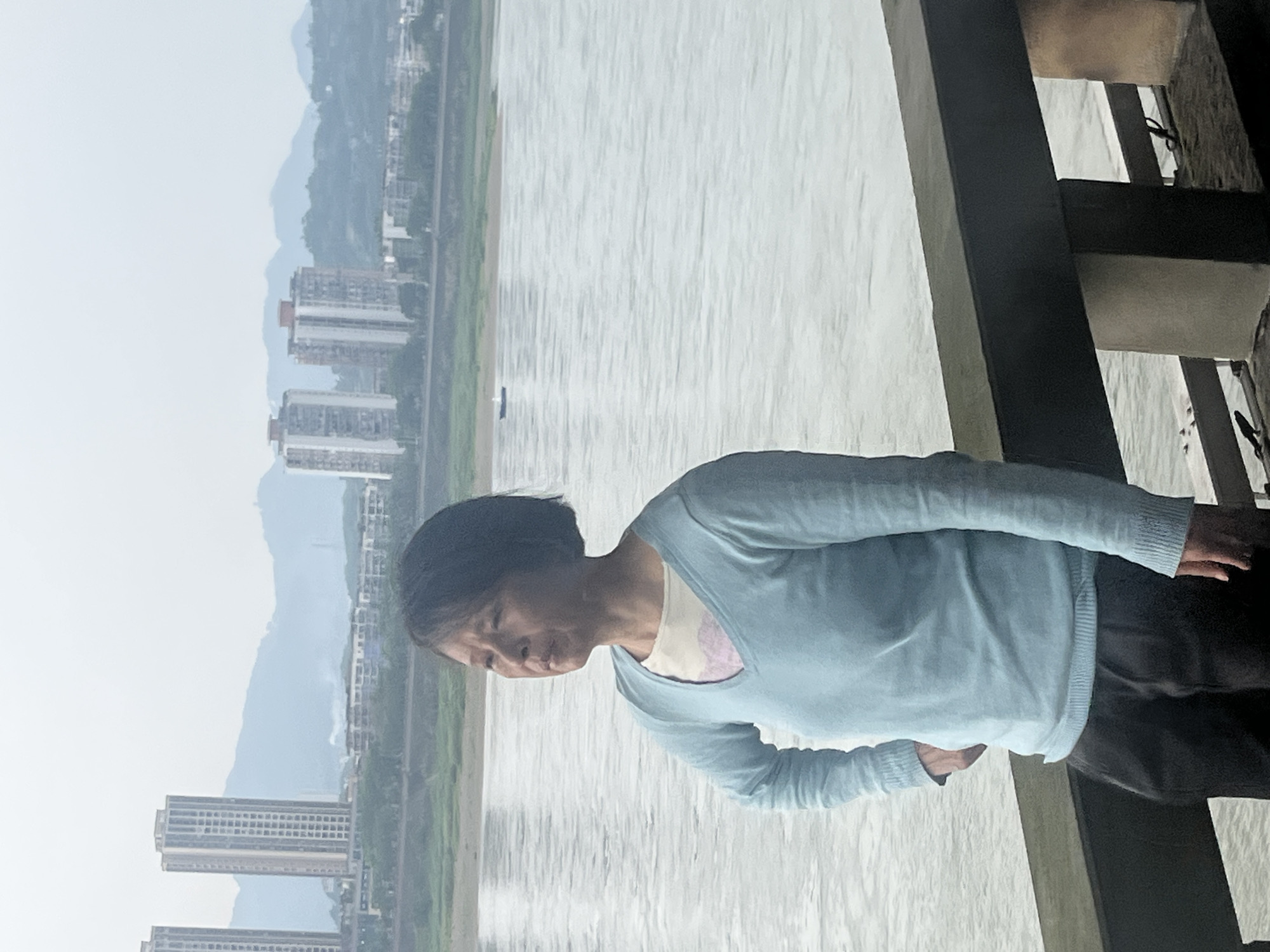 An Asian woman stands on a wooden structure with metal cables overlooking a wide, light gray river. The woman has dark hair and is wearing a light blue sweater over a light-colored top and dark pants. In the background, light gray buildings rise along the riverbank near distant, hazy mountains. A small boat is visible on the river. This scene appears to be taken from Jiangjin, China.