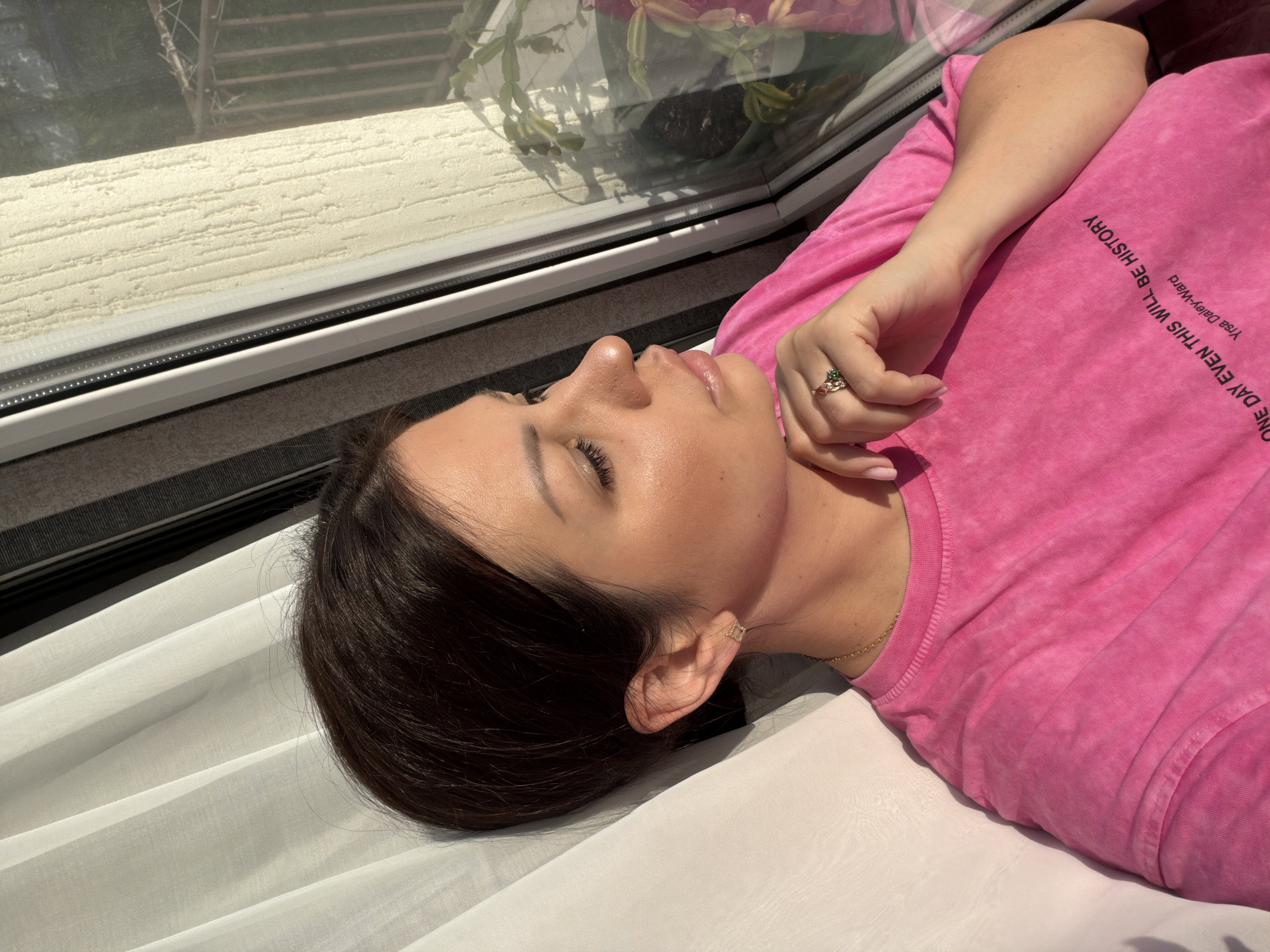 The image shows a woman with dark hair, wearing a bright pink t-shirt with text that reads "ONE DAY EVEN THIS WILL BE HISTORY" and "Yrsa Daley-Ward". She is wearing a gold earring and a ring on her right hand. She is looking out a window, with a curtain on the left side and the window frame and textured wall on the right. The environment suggests a residential interior with sunlight streaming in. The location is identified as Sochi, Russia.