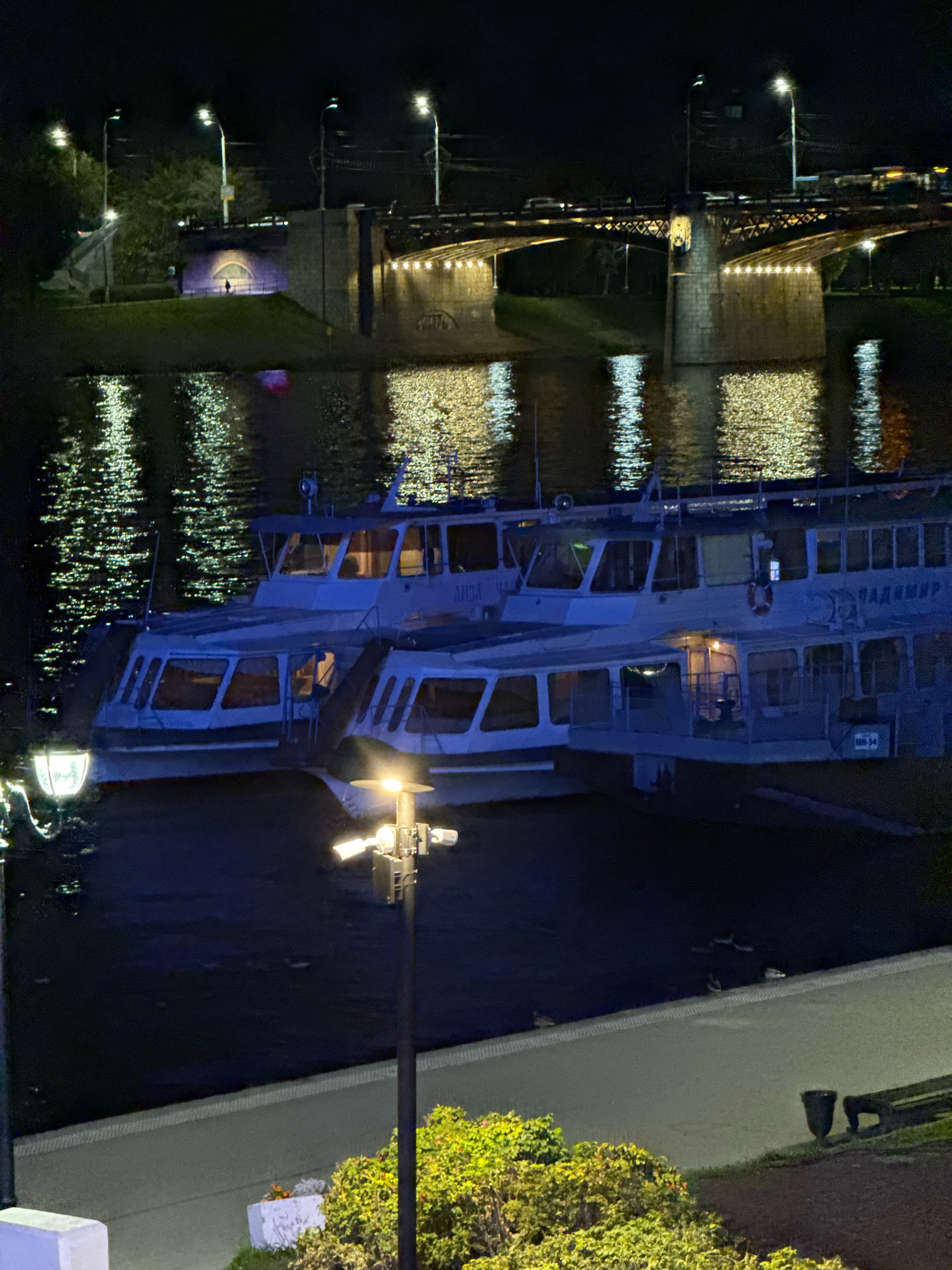The image, taken at night in Tver, Russia, shows two pleasure boats docked on a body of water, likely the Volga River. The boats are illuminated from within and by external lights. In the background, a bridge spans the river, with lights visible on its structure and on the roadway above, where cars are moving. The water reflects the lights from the bridge and the sky, creating a shimmering effect. A single person is visible walking near an illuminated archway on the embankment. Streetlights are present along the riverbank, and foliage can be seen in the foreground. The scene depicts a nighttime urban landscape with a focus on the illuminated river, boats, and bridge.