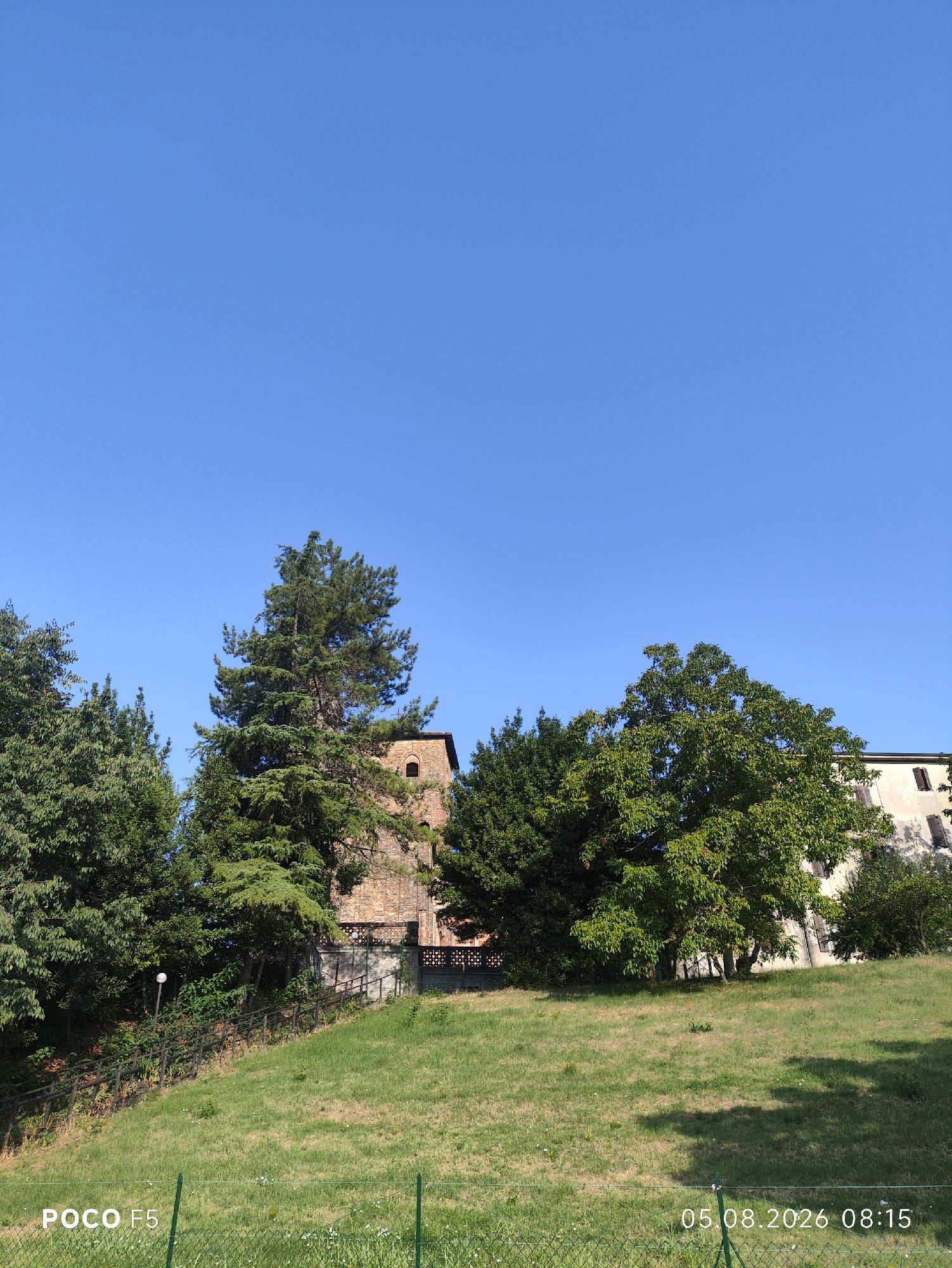 The image captures a sunny day in the city of Collecchio, Italy. In the foreground, a neatly mown grassy slope descends from right to left. A green metal fence runs along the bottom of the frame, with the text "POCO F5" visible on the left and "05.08.2026 08:15" on the right. The middle ground features a variety of trees, including a tall conifer and a broadleaf tree with dense foliage. Behind the trees, a rustic brick building with arched windows can be seen, suggesting it might be an old tower or part of a historical structure. A dark wooden fence borders the property in the middle ground. The background is dominated by a clear, bright blue sky, indicating fair weather. The scene appears to be a park or a residential area with ample greenery. There are no people visible in the image.