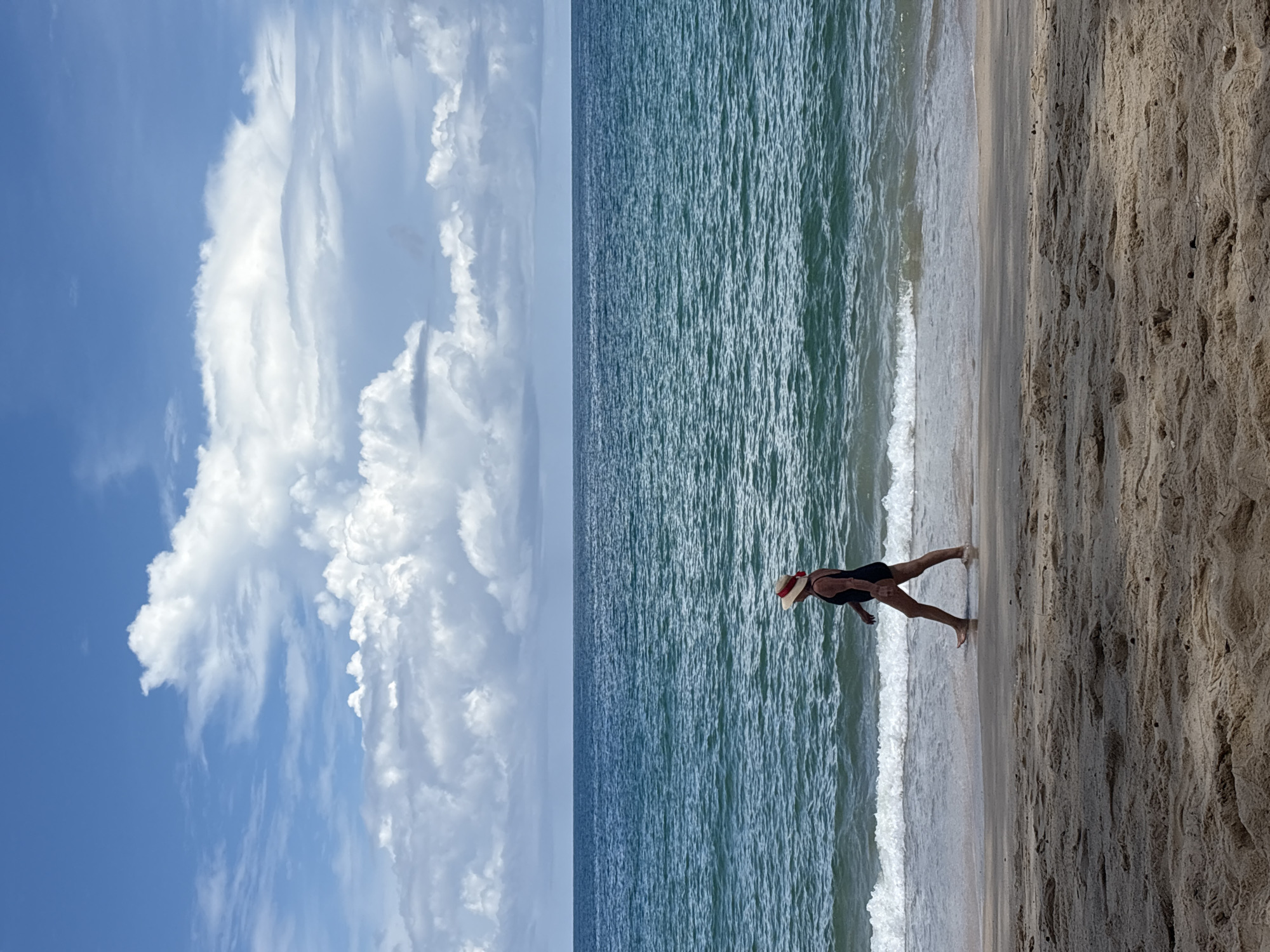 A person wearing a black swimsuit and a straw hat with a red ribbon walks along the shoreline of a sandy beach. The person is facing left in profile. The sea, with calm waves, is to the person's left, and the sandy beach is to the person's right. The sky above is blue with white, fluffy clouds. The location is the beach in Choeng Thale, Thailand.