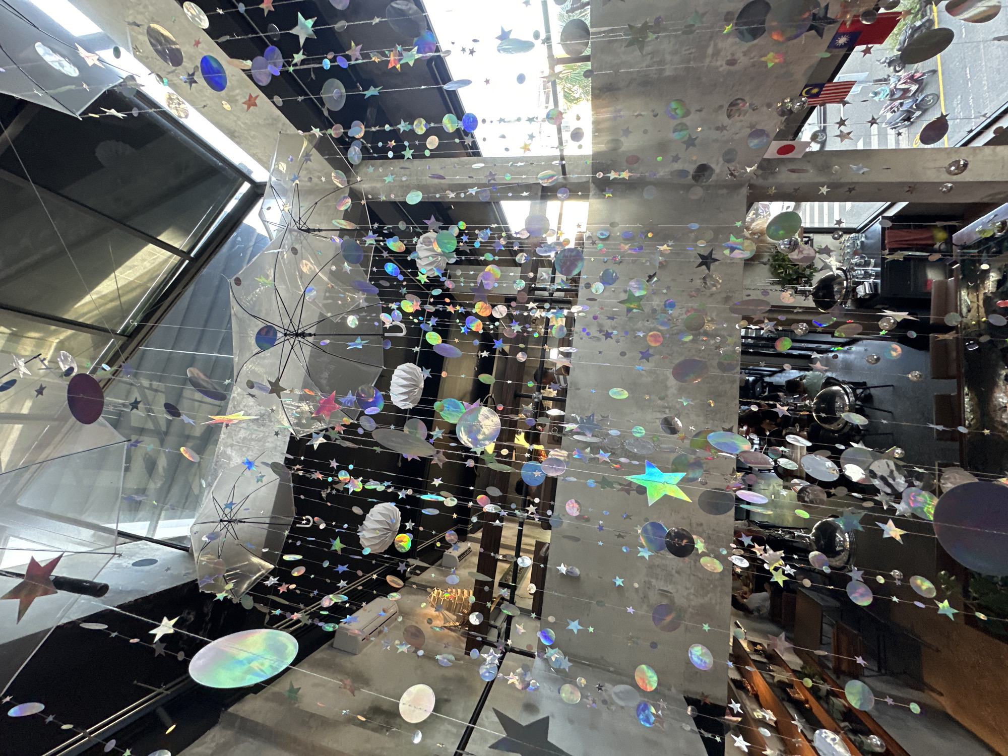 The image depicts the interior of a cafe in Đà Nẵng, Vietnam, with a focus on a unique ceiling decoration. Hanging from the ceiling are numerous clear umbrellas, each adorned with strings of iridescent star and circular cutouts that catch the light. These shimmering ornaments create a whimsical, celestial effect. Below the umbrellas, more strings of these cutouts hang down, creating a layered, curtain-like appearance. The cafe's interior features exposed concrete walls and multiple levels, suggesting an industrial-chic design. In the background, patrons can be seen seated at tables, with some activity visible on the street outside, including parked motorcycles and passing vehicles. Flags are displayed on a pillar, including the Japanese flag, the Malaysian flag, and the Taiwanese flag, indicating a potentially international clientele or theme. The overall atmosphere appears to be a bright and airy space, with sunlight streaming through the windows.