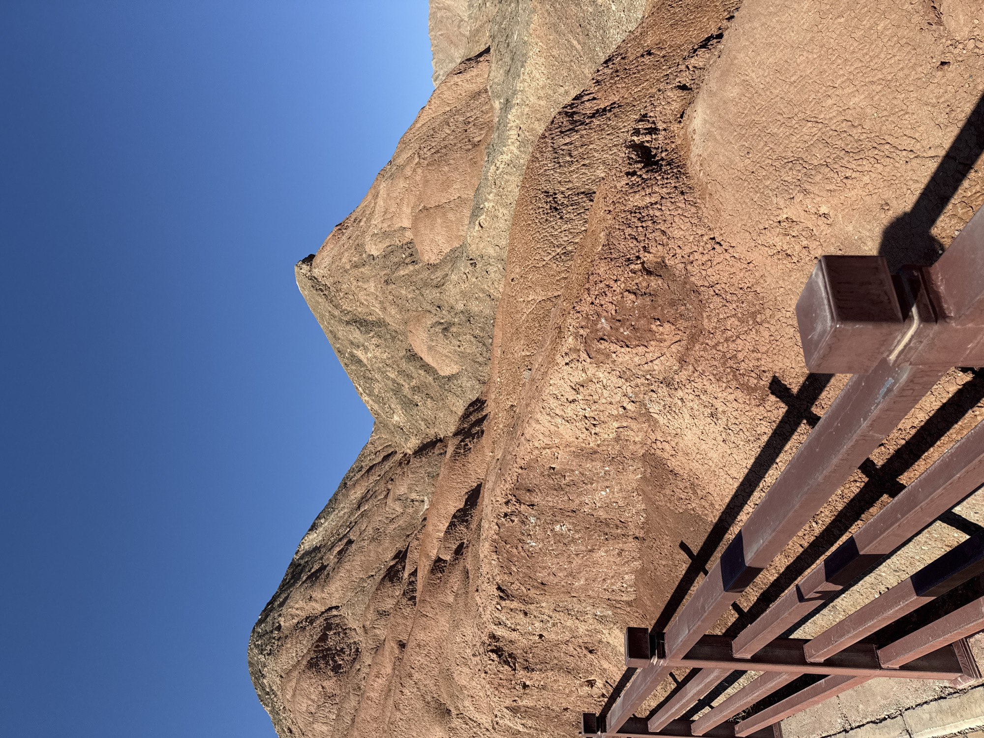 The image presents a dramatic natural landscape, featuring immense rock formations under a clear, vibrant blue sky. The scene is dominated by towering, reddish-brown geological structures that exhibit varied textures and stratification, suggesting ancient erosion and distinct geological layers. The rock surfaces range from finely cracked and sandy, almost desert-like ground, to rugged, undulating cliff faces marked by shadows that emphasize their three-dimensional nature.
In the foreground and extending into the middle ground on the right, a dark brown metal railing or part of a viewing platform is visible. This man-made structure is sturdy and casts long, sharp shadows onto the textured ground and lower rock sections, indicating bright, direct sunlight. No people are visible, allowing the expansive natural environment to take center stage.
The time of day is clearly sunny daytime, characterized by the cloudless, deep blue sky and the crisp, well-defined shadows cast by both the railing and the contours of the rock formations. The weather appears clear and calm. While no specific activities are depicted, the presence of the railing suggests a designated area for observation, possibly a scenic overlook or trail, within a natural park or attraction. This type of rugged, richly colored rock landscape is consistent with certain geological regions, such as those found near Nantai, China. There is no visible text in the image. The overall impression is one of majestic natural beauty and geological grandeur.