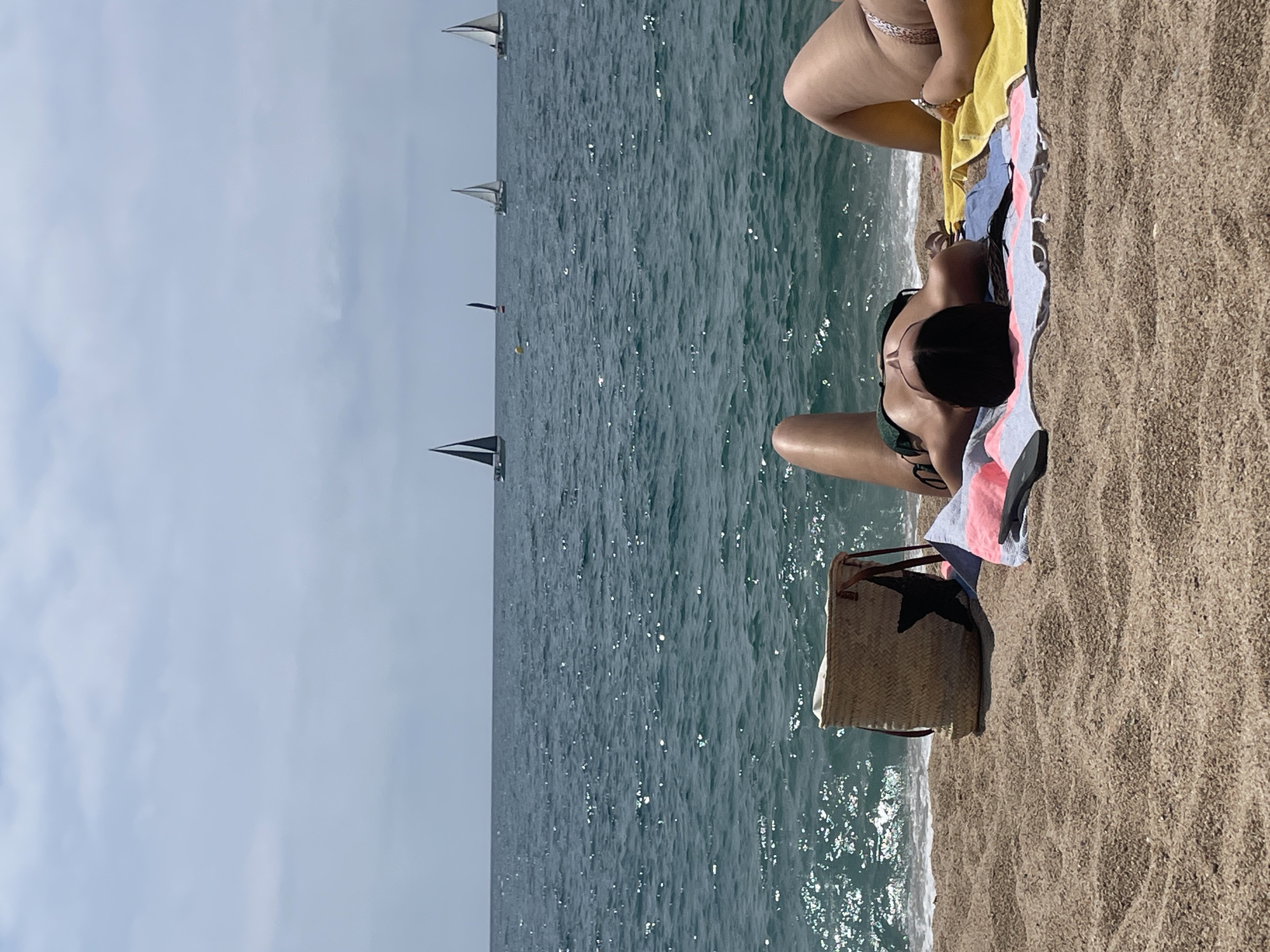 This image depicts a sunny day at the beach in Canet de Mar, Spain. Two women are lying on towels on the sand, facing away from the camera. The woman in the foreground is wearing a dark green bikini and has her head resting on her arms. The woman in the background is wearing a patterned bikini and is also lying on a towel. A woven straw bag with a black star design sits on the sand near the woman in the foreground. The sea is visible behind them, with several sailboats in the distance. The sky is overcast.