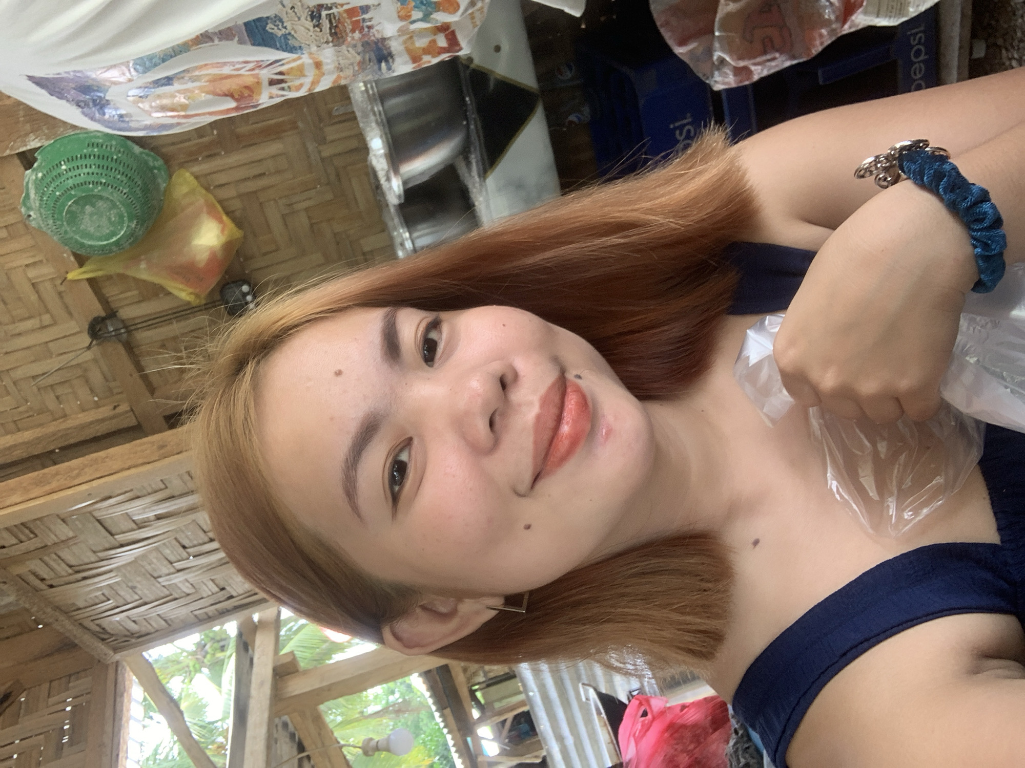 A close-up, selfie-style shot features a young woman with light brown hair, an open-mouthed smile, and subtle makeup. She is wearing a dark blue sleeveless top and a silver square earring in her left ear. She clutches a clear plastic bag in her right hand and sports a blue scrunchie with charms on her left wrist. The background is an outdoor, rustic setting, possibly a market or roadside eatery in Kiblawan, Philippines. The structures are made of natural materials like bamboo and woven palm leaves. In the background, various items are visible, including a green plastic colander hanging on the wall, a yellow plastic bag, and metal containers. Sunlight filters through the thatched roof, illuminating the scene.