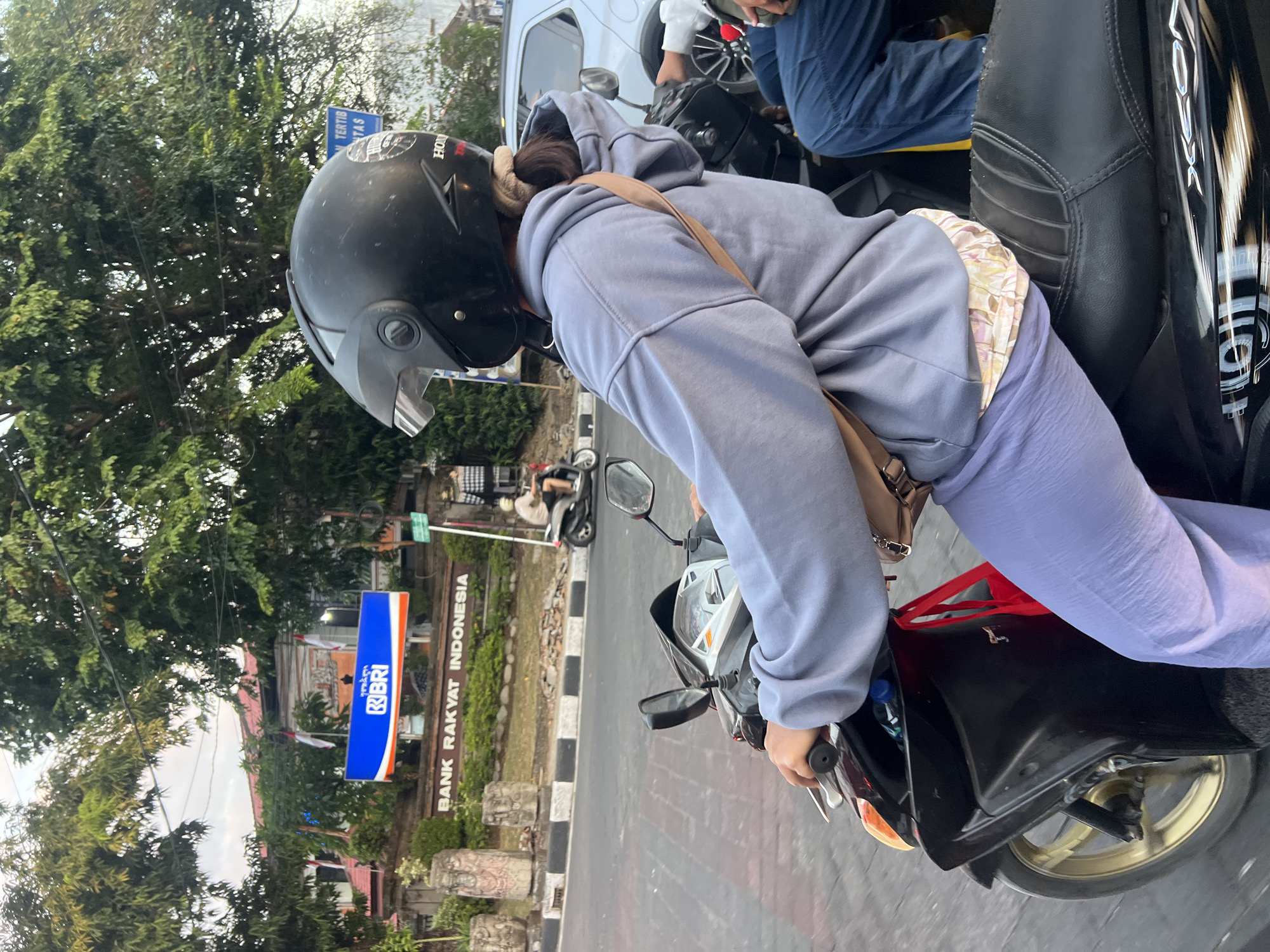A person wearing a black helmet and a light blue hooded sweatshirt is riding a black motorcycle on a street in Denpasar, Indonesia. The person is wearing light purple pants and a patterned garment is visible underneath. A brown strap bag is slung across the person's chest. In the background, a sign for "BANK RAKYAT INDONESIA" is visible, along with trees and other vehicles. The sky appears overcast, suggesting it might be late afternoon or early morning. The road surface is paved, and there are some decorative stone pillars on the side of the road. Another motorcycle with a rider is visible further down the street.