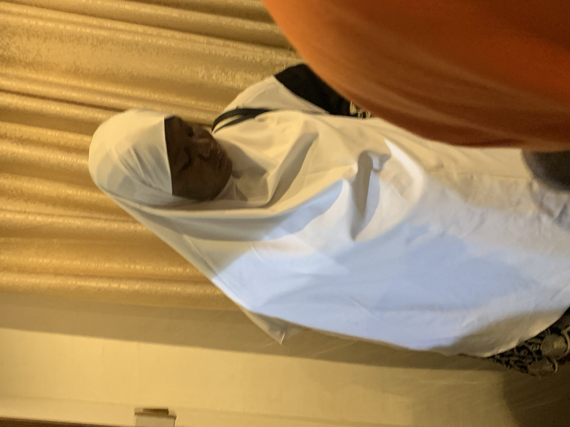 A dark-skinned person lies under a white sheet and head covering. The background shows golden curtains. The location is Jos, Nigeria.