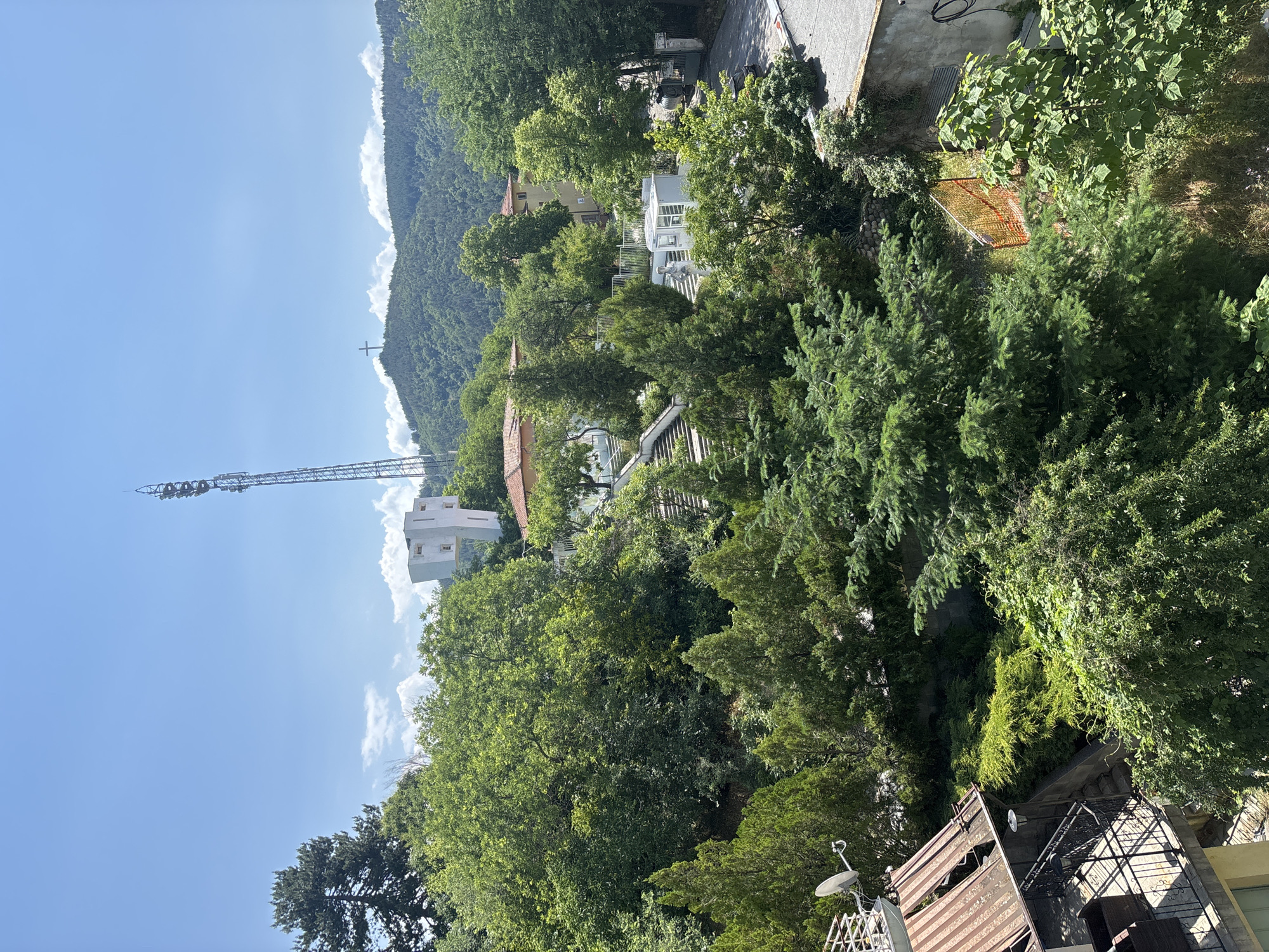 This media file captures a wide, elevated view of a mountainous landscape, likely depicting a section of Blagoevgrad, Bulgaria, under bright daylight. The scene is dominated by lush, dense green trees covering the hillsides and surrounding residential structures. The sky is a clear, vibrant blue with scattered white, fluffy clouds, suggesting fair weather.
Prominently featured on the left side of the image is a tall, grey metal telecommunications tower, extending upwards into the sky. On a distant mountain peak or ridge, a large cross is visible, partially obscured by clouds, serving as a distinct landmark.
Nestled among the trees are several buildings, mostly residential, characterized by light-colored facades and some with terracotta-tiled roofs. In the foreground and lower right, details such as a rusty corrugated metal roof, a balcony with a dark metal railing, and outdoor furniture (table and chairs) are visible, hinting at human presence. Crops reveal further details, including a white, greenhouse-like structure or gazebo with a small statue, and a section of orange mesh netting or fencing amidst the vegetation.
No individuals are visible, and no specific activities or events are taking place, rendering a tranquil and expansive overview of a town integrated into a natural, forested mountainous environment. There is no visible text in the image.