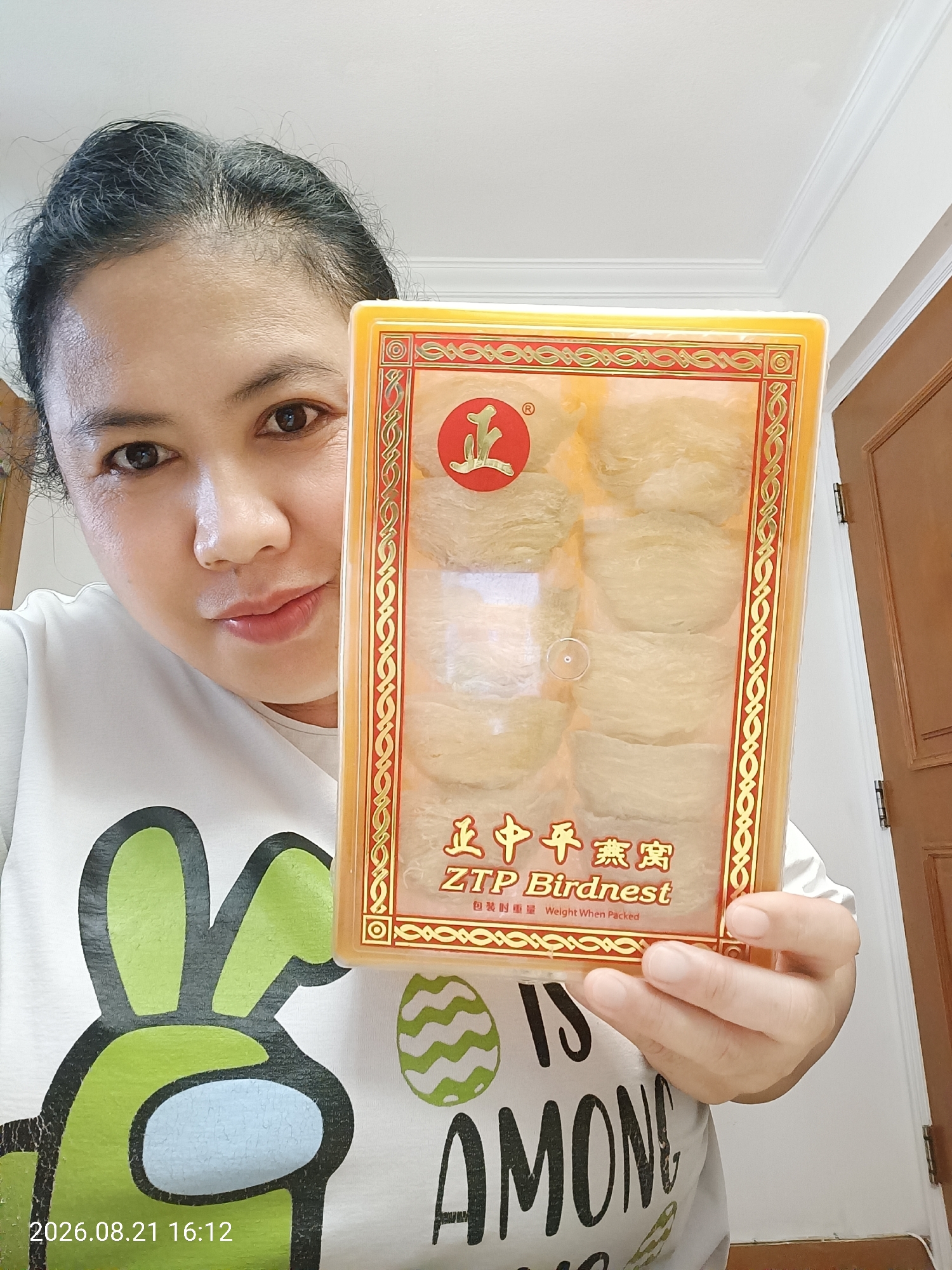 A medium shot captures a woman of Asian descent holding a box of bird's nest products. She is positioned in the foreground, with her head and shoulders visible. The woman has dark hair, brown eyes, and fair skin. She is wearing a white t-shirt with a green character graphic, possibly from the game Among Us, and the text "IS AMONG" printed below it. The bird's nest box is rectangular with a clear plastic lid, revealing several bird's nest products inside. The box has decorative golden and red patterns and text in Chinese characters and English. The background consists of a white ceiling with molding, a beige wall with a wooden door, and a hint of a doorway. The lighting suggests an indoor setting, possibly a home. A timestamp overlay in the bottom left corner of the image reads "2026.08.21 16:12". The image appears to be a selfie. The location is not specified in the image.