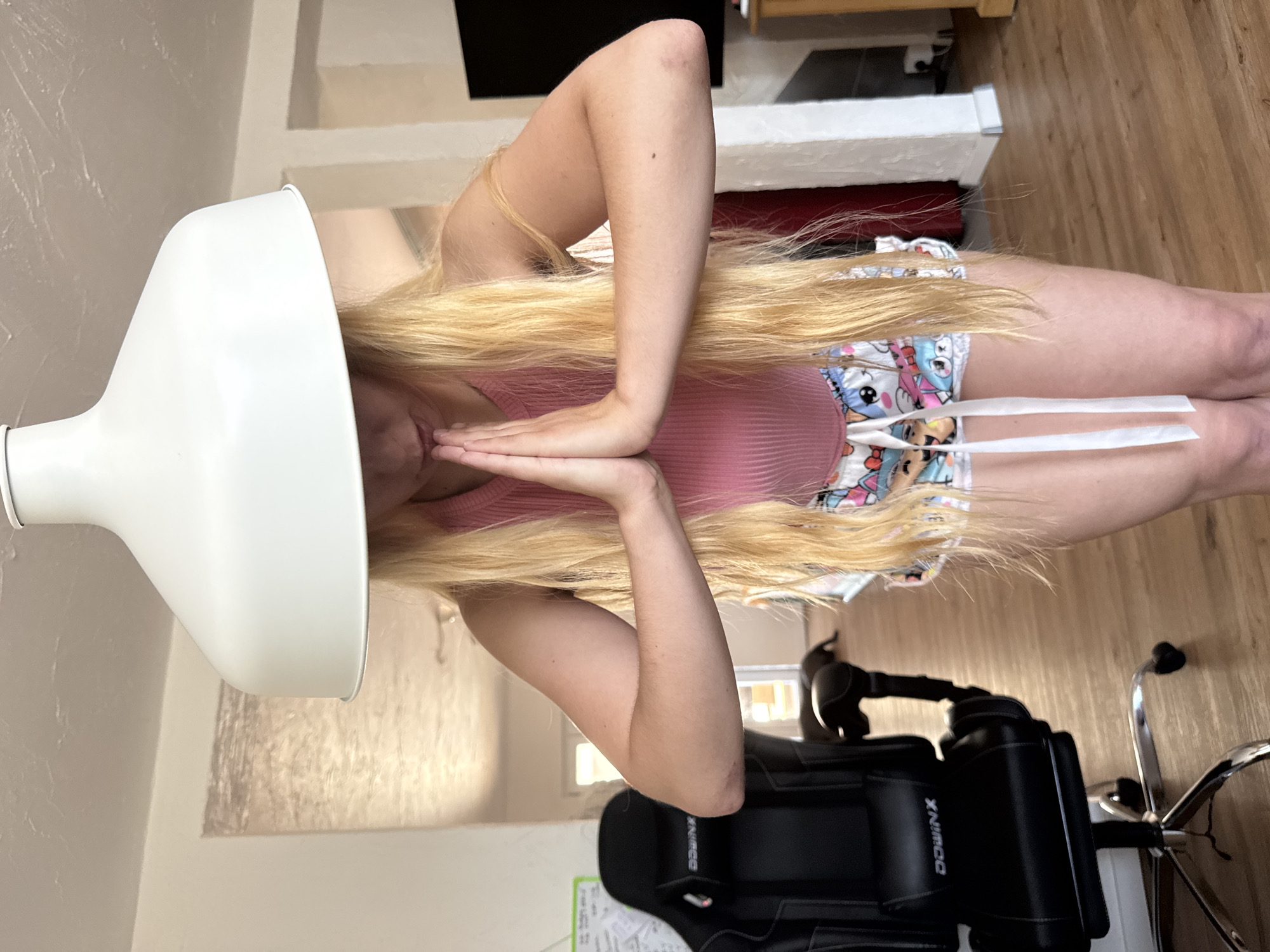 The image shows a person with long blonde hair, wearing a pink tank top and patterned shorts. Their hands are clasped together in front of their face, and they appear to be under a white hair dryer. The setting appears to be indoors, possibly a home, with wooden flooring visible and what looks like a chair in the background. The lighting suggests it is daytime.