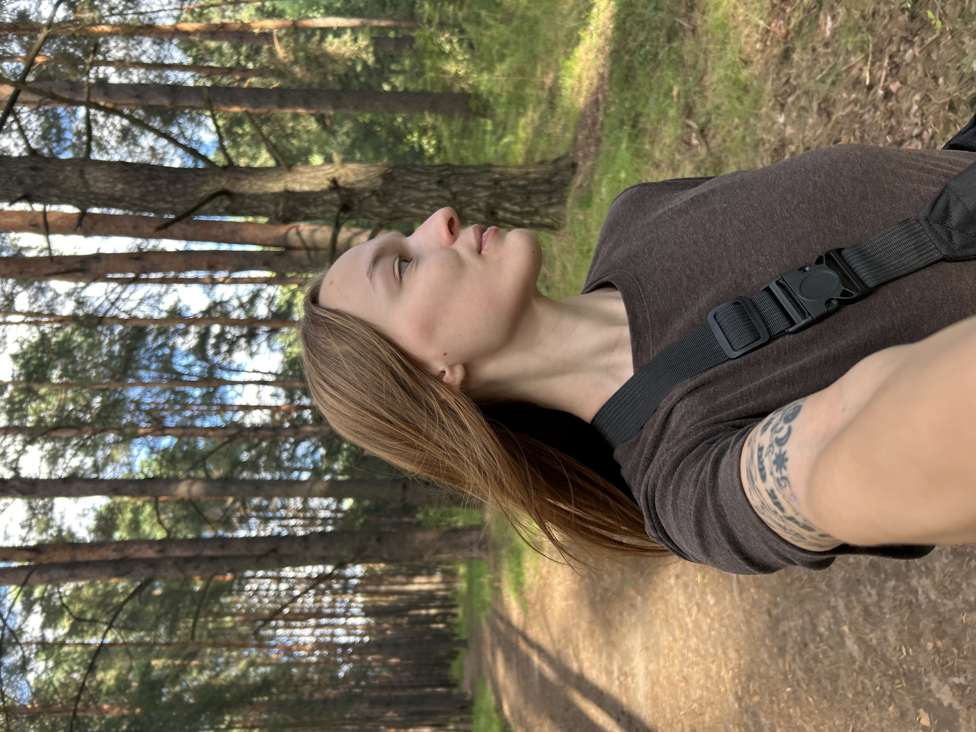 A young woman with shoulder-length brown hair is captured in a selfie-style photograph, positioned in the right half of the frame and facing to her left. She wears a dark brown t-shirt with a black strap across her chest, likely part of a bag. Her left arm is extended, showcasing a tattoo band around her bicep with various symbols and text.
The background consists of a sun-dappled forest path, leading into a dense pine forest. Tall, slender pine trees dominate the scene, with a lush green undergrowth of grass and foliage. The sky is visible through the trees, appearing blue with white clouds. The lighting suggests it is daytime, with sunlight casting shadows on the path. The image appears to be taken in a natural outdoor environment, possibly a park or forest trail, consistent with the landscape found in and around Chaikovsky, Russia, known for its extensive forested areas and recreational paths.