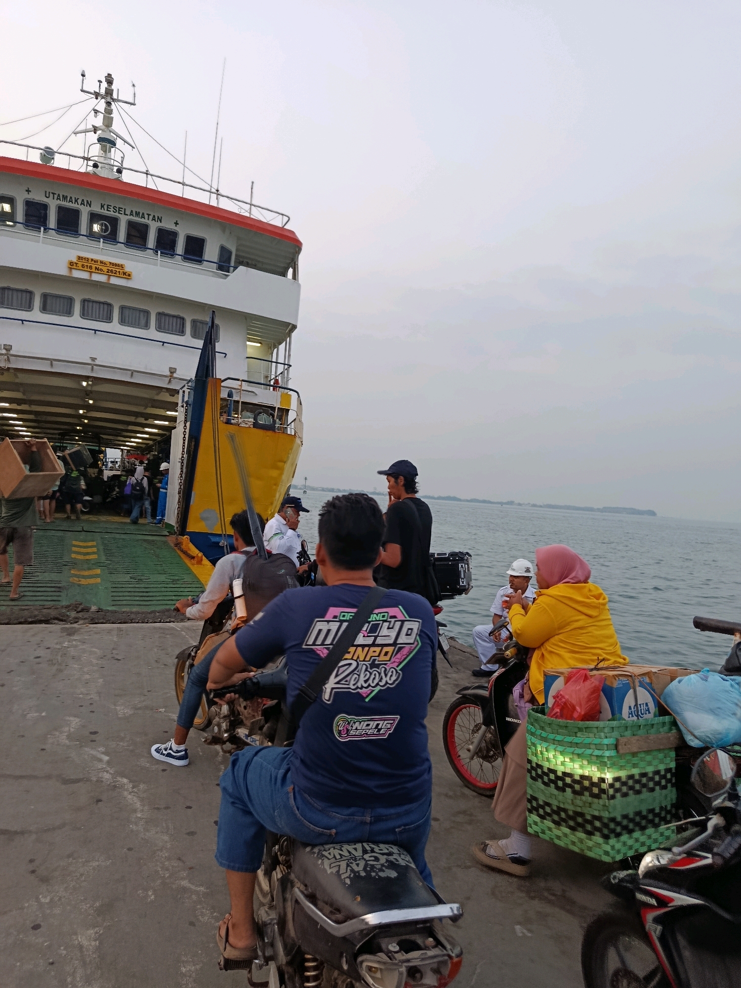 The image depicts a ferry loading scene at a port, likely in Jepara, Indonesia, given the context. In the foreground, several individuals are on motorcycles, preparing to board or having just boarded the ferry. The ferry, a large vessel with multiple decks and a ramp open for vehicle access, dominates the background. The sky is overcast, and the sea is visible to the right.
In the immediate foreground, a person wearing a blue t-shirt with text and blue jeans is seated on a motorcycle. To their right, another motorcycle is visible, carrying a passenger in a yellow hoodie and a pink headscarf. This passenger is seated and appears to be holding something to their mouth. Behind them, a person in a white uniform and cap is also seated on a motorcycle. Further to the left, more people are seen, some with backpacks, and a person is carrying a cardboard box. The ferry's deck is a sloped ramp with yellow markings.
The ferry itself is white with a red stripe along the top deck and signage that reads "UTAMAKAN KESELAMATAN" (Prioritize Safety). A yellow section is visible on the side of the ferry, and a blue flag or banner is partially unfurled. Essential details like ship identification numbers are present on a yellow plaque. The environment appears to be a port with concrete ground and a body of water to the right. The lighting suggests it is either early morning or late afternoon.