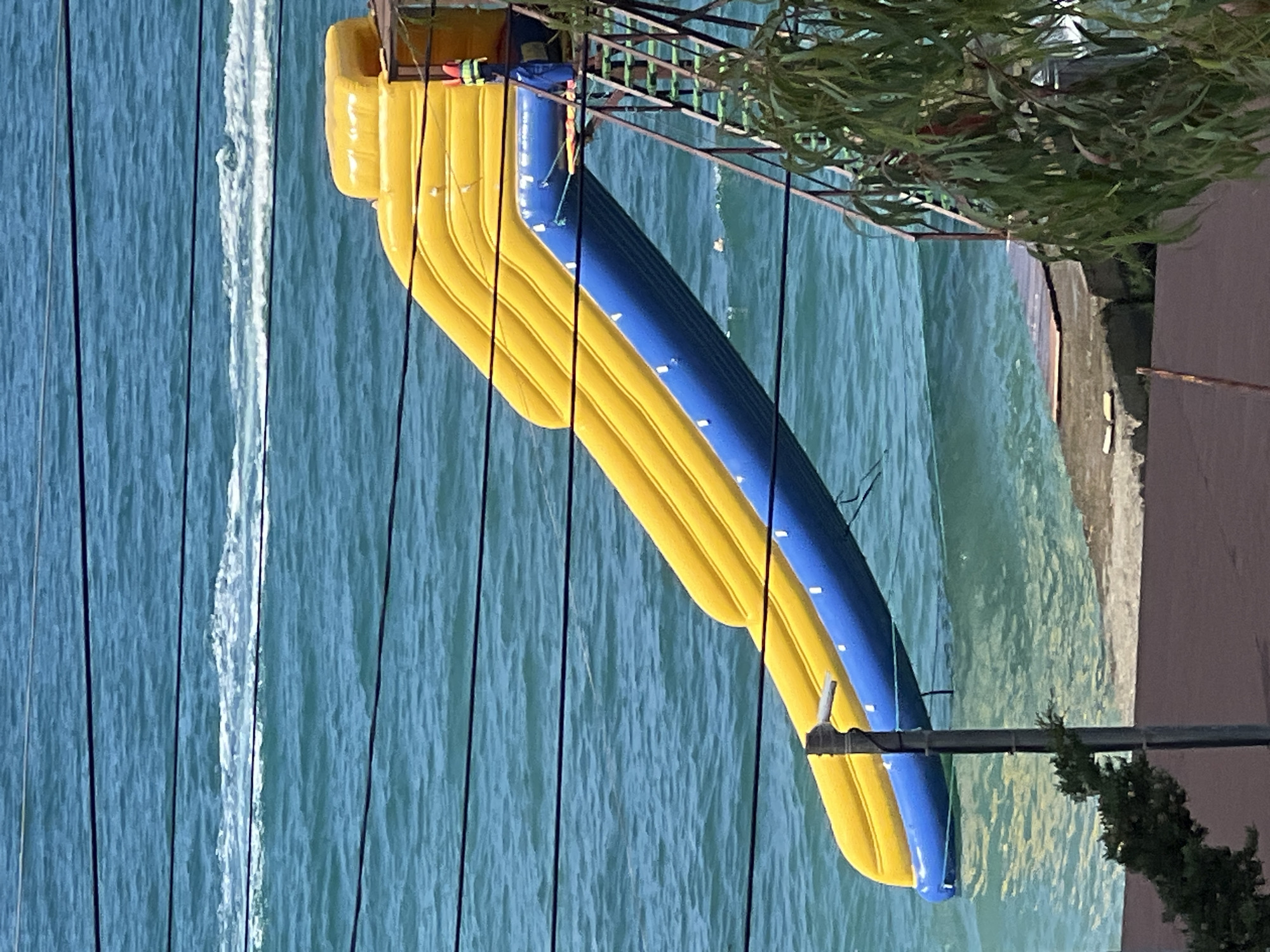 This is a vertical, full shot of an inflatable water slide, likely at a beach or water park, with the turquoise water of the sea in Sochi, Russia, visible in the background. The slide is predominantly yellow and blue, with a yellow inflatable entrance at the top and a long blue slide section. A metal structure supporting the slide is also visible, along with some green foliage from trees overhanging the scene. A person wearing a life jacket is partially visible at the top of the slide, suggesting preparation for or the end of an activity. The water appears choppy, indicating it might be a windy day. The lighting suggests it is daytime, with bright sunlight reflecting off the water. The overall impression is of a recreational setting on a warm day.