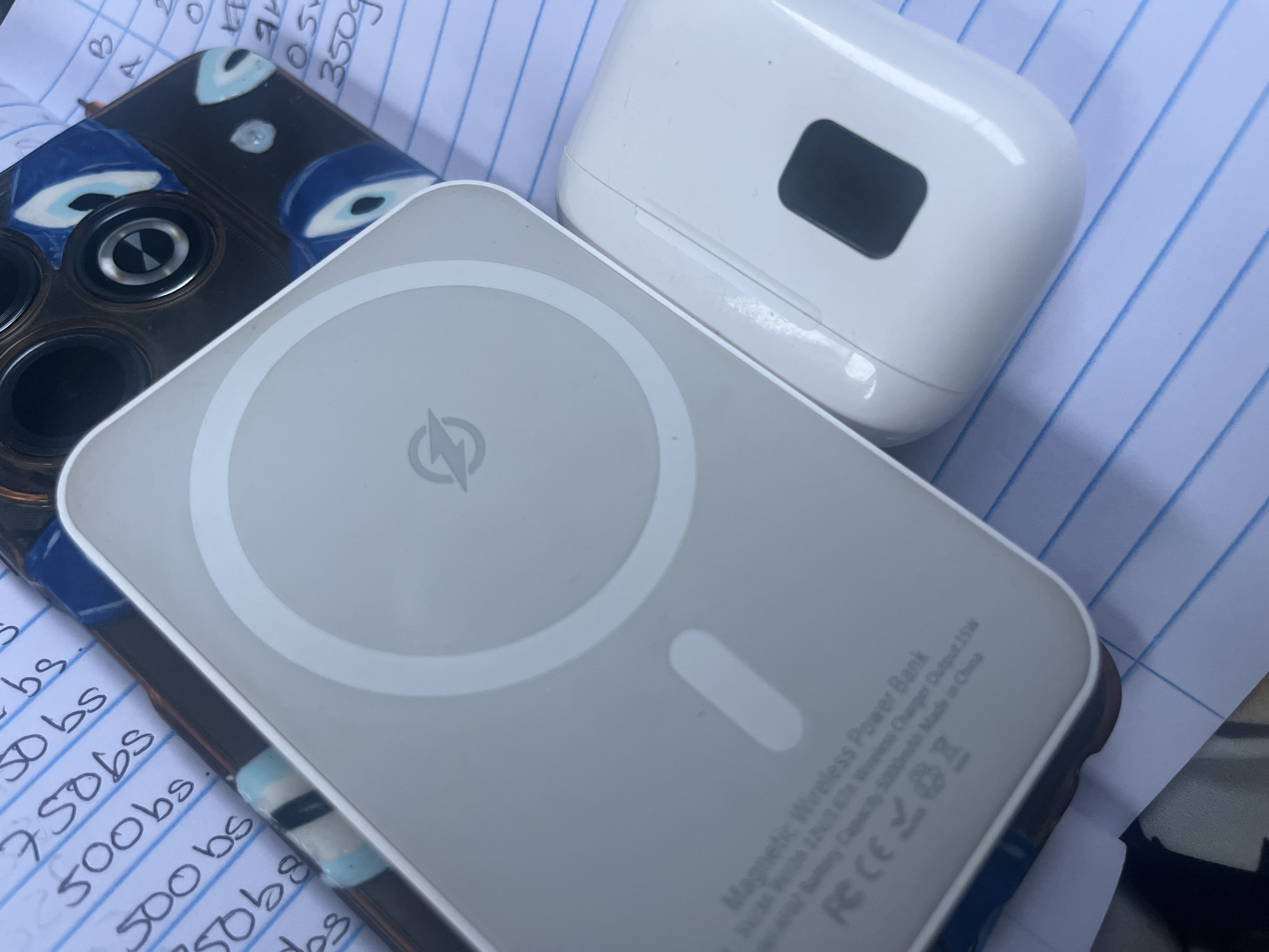 This is a close-up, overhead shot of a white magnetic wireless power bank resting on a blue-lined notebook. The power bank has a circular indentation on its surface with a lightning bolt logo in the center. To the right of the power bank, a white earbud charging case is visible. Partially in the background, a phone case with an evil eye design can be seen. The notebook pages appear to have handwritten notes, including numbers and "bs." The overall scene suggests a personal item setting, possibly a desk or table. The location in Conuco Viejo, Venezuela is not visually represented in this image.