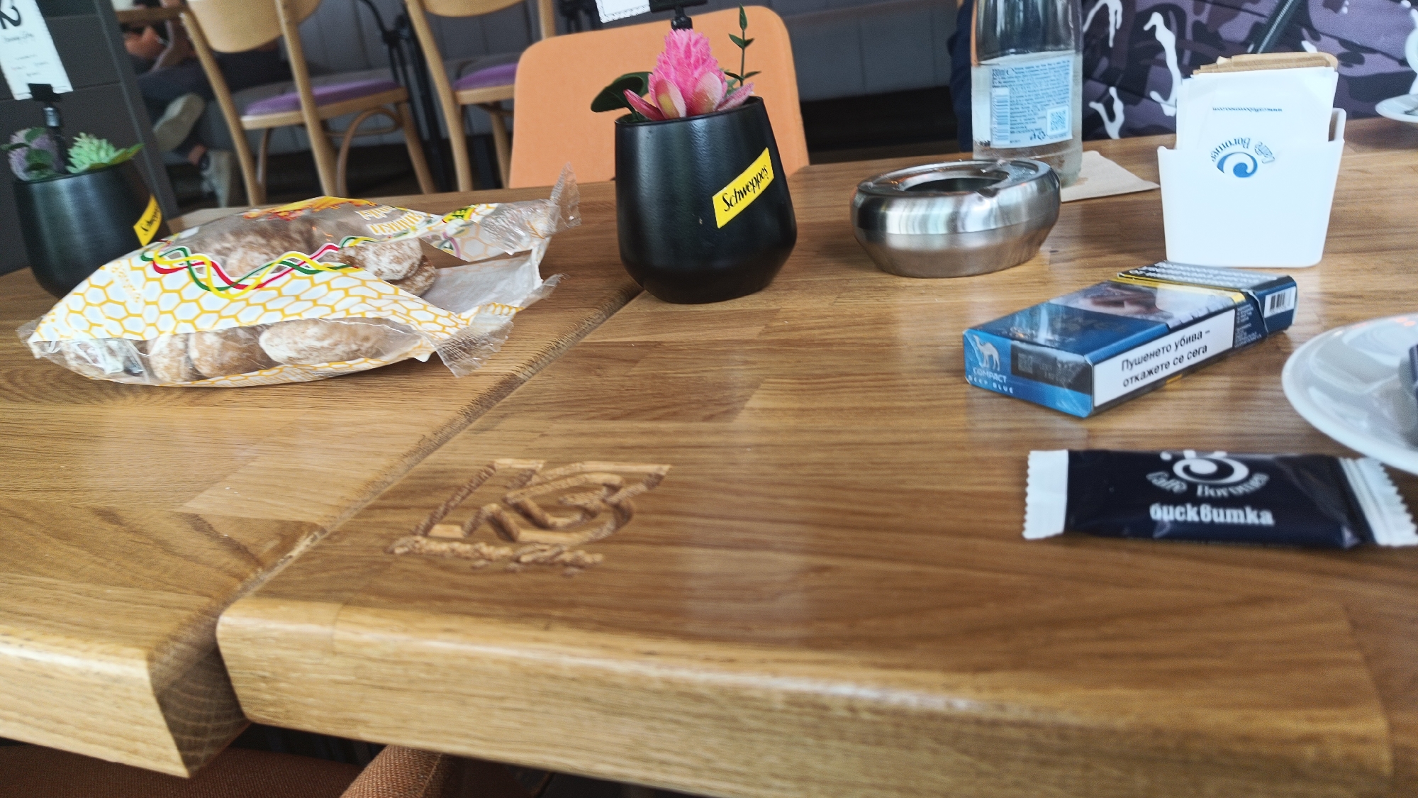 The image depicts a tabletop scene at a café in Burgas, Bulgaria. The table surface is made of light-colored wood. The following objects are present: a partially consumed bag of cookies, a black flower pot with a pink flower and a "Schweppes" sticker, an ashtray, a pack of "Camel" brand cigarettes with text in Bulgarian, a sugar packet with Cyrillic writing, a white tissue dispenser with a wave design, a water bottle, and a saucer with a spoon. People can be seen in the background.